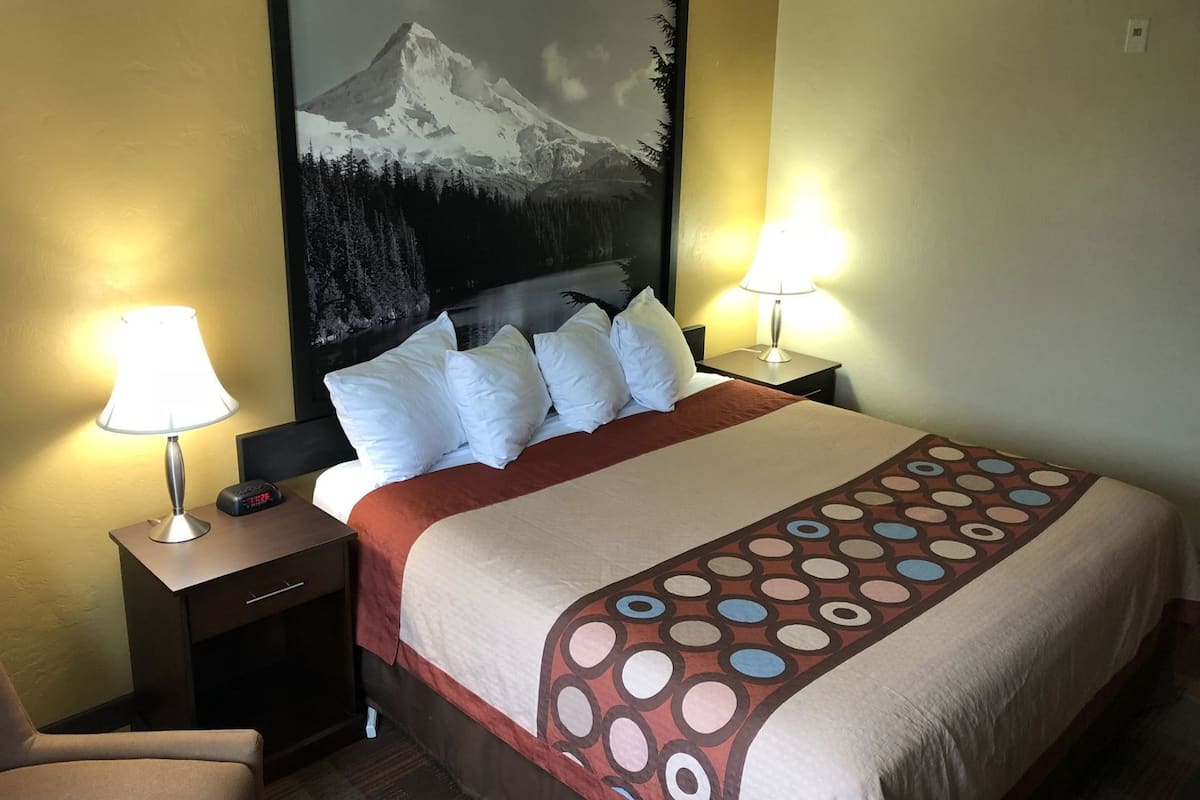 A hotel room with a bed, two bedside lamps, a nightstand, a chair, and a large mountain painting on the wall.