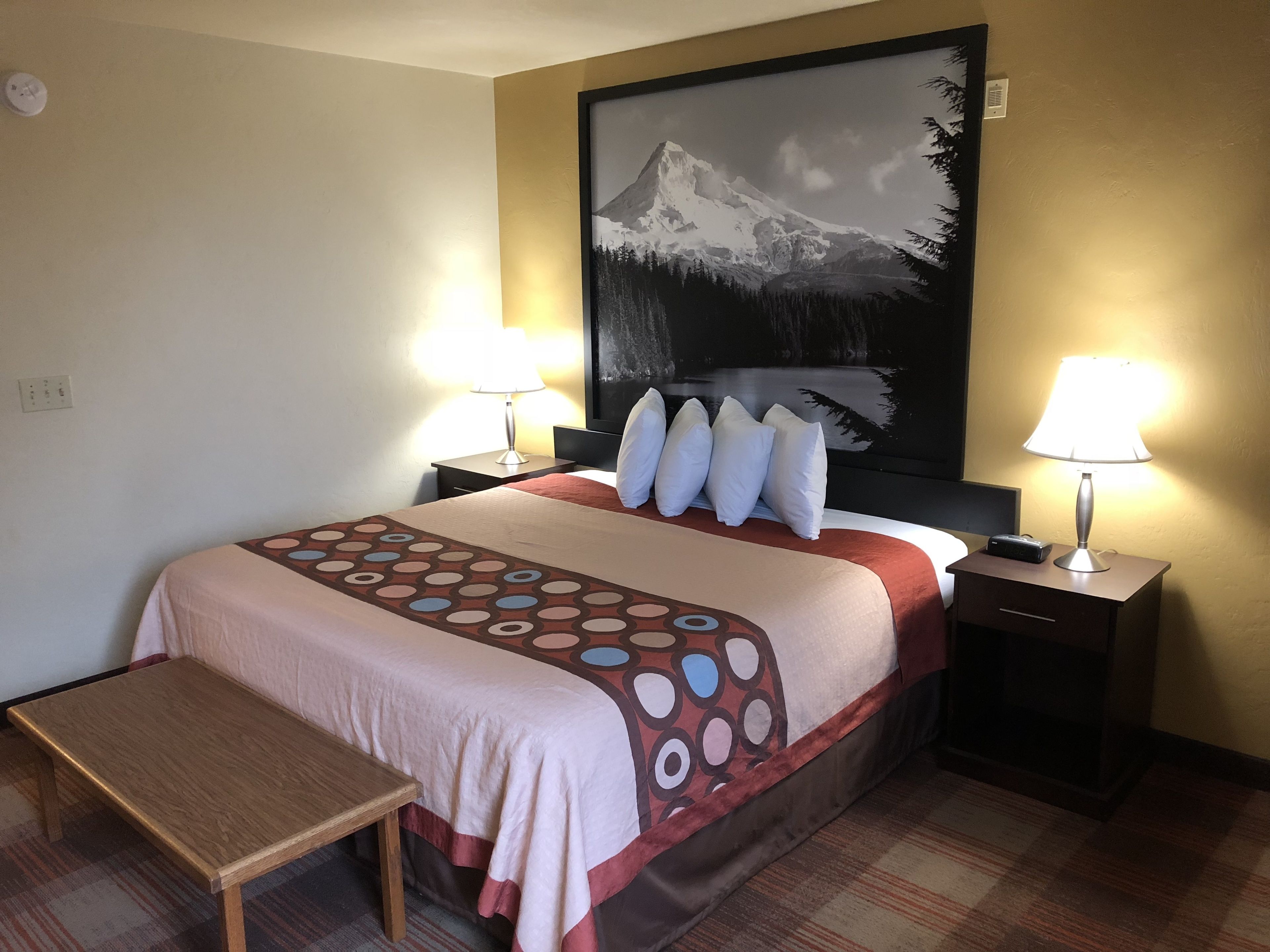 A hotel room with a bed, two bedside tables, a nightstand, a bench, and a large painting on the wall.