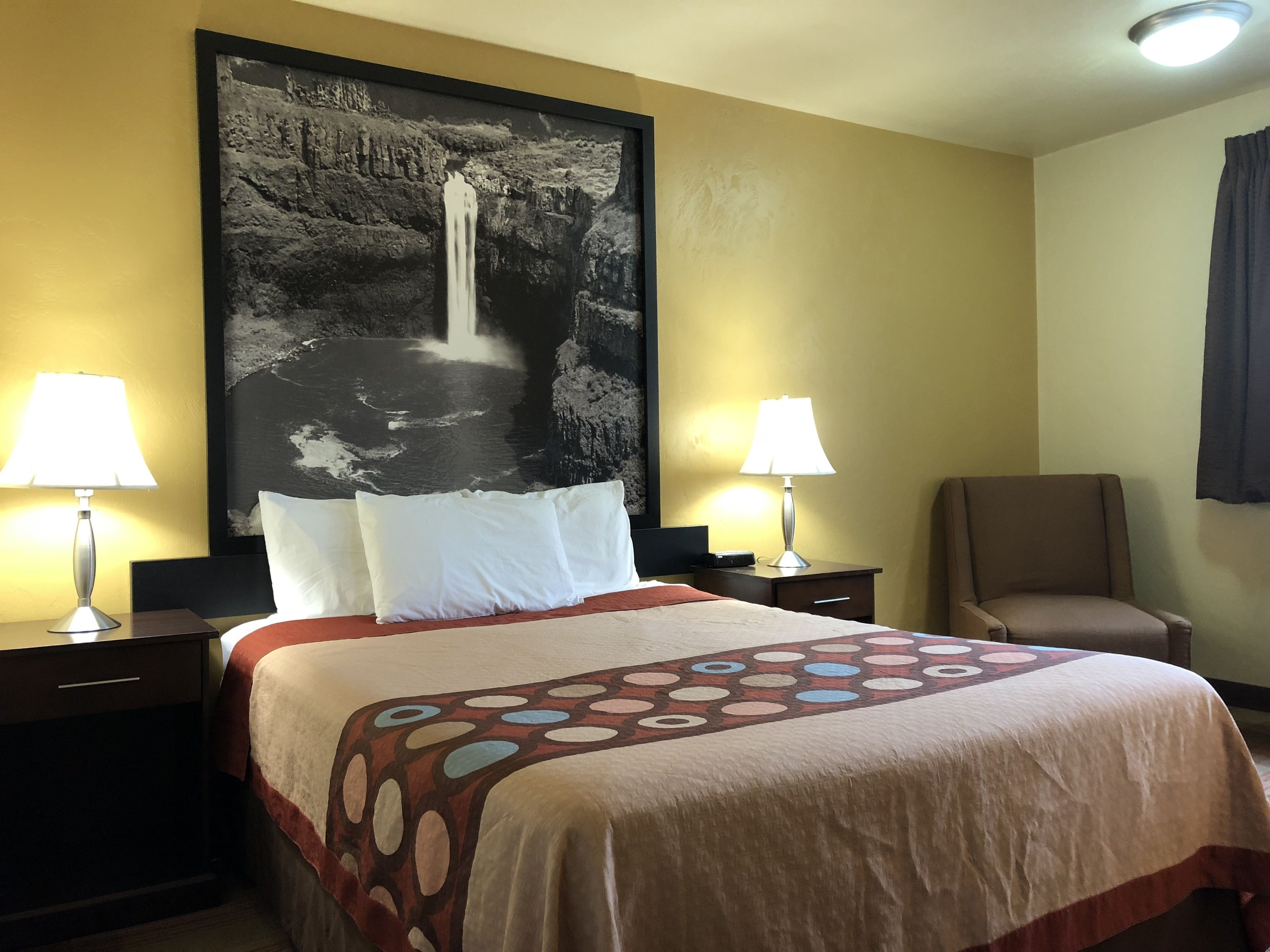 A hotel room with a bed, two bedside lamps, a chair, and a large wall painting of a waterfall.
