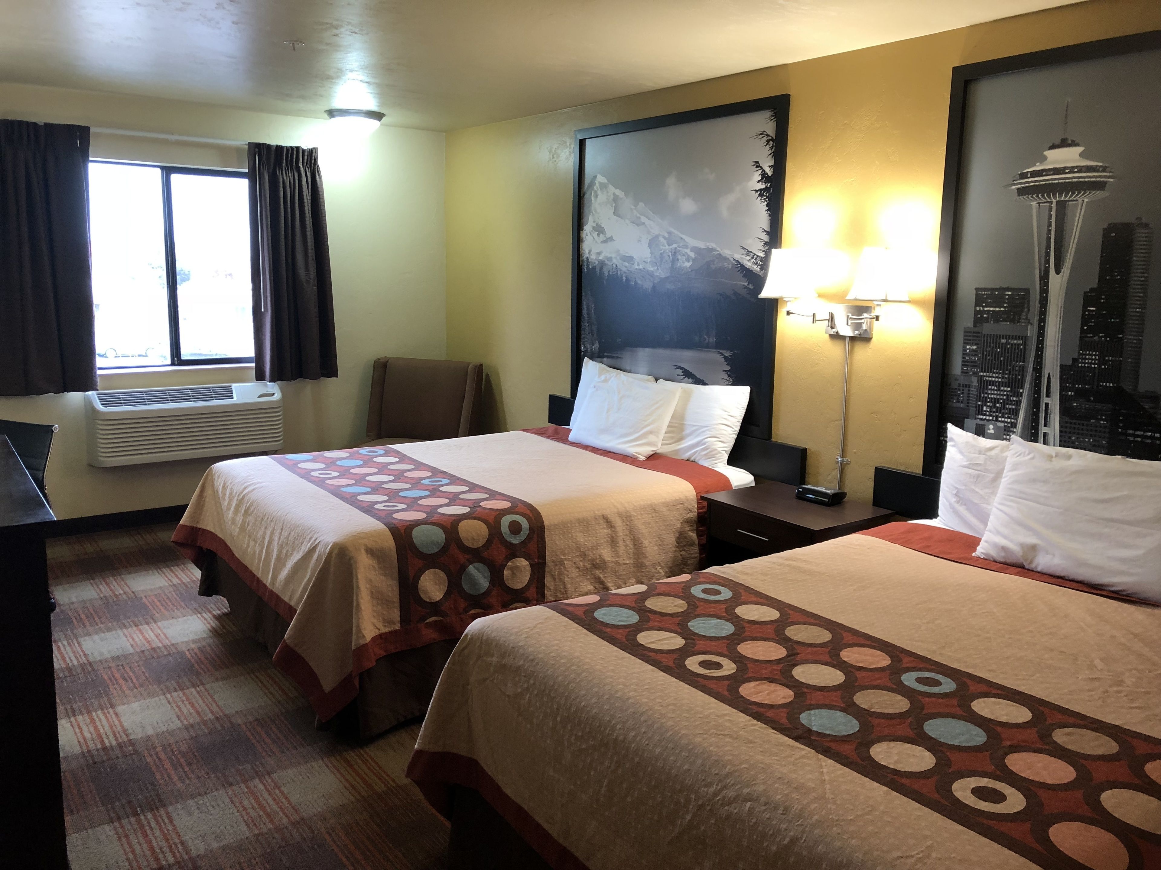 Habitación de hotel con dos camas, un ventanal con cortinas, aire acondicionado de pared, una silla, una mesita de noche con lámpara y vistas a la ciudad.
