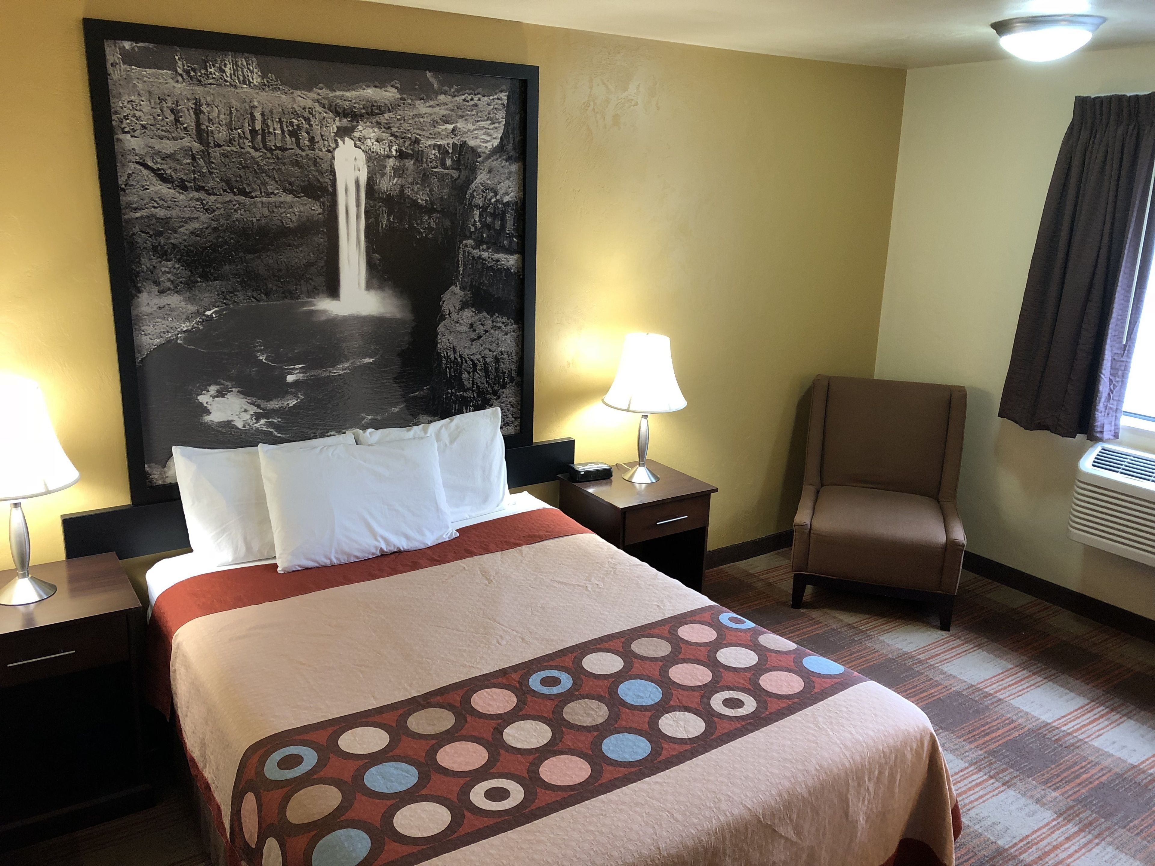 Une chambre d’hôtel comprenant un lit, deux tables de chevet, une chaise, un grand tableau mural et une fenêtre avec des rideaux.