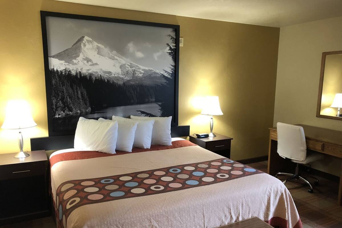 A hotel room with a bed, two bedside lamps, a desk with a chair, and a large mountain painting on the wall.