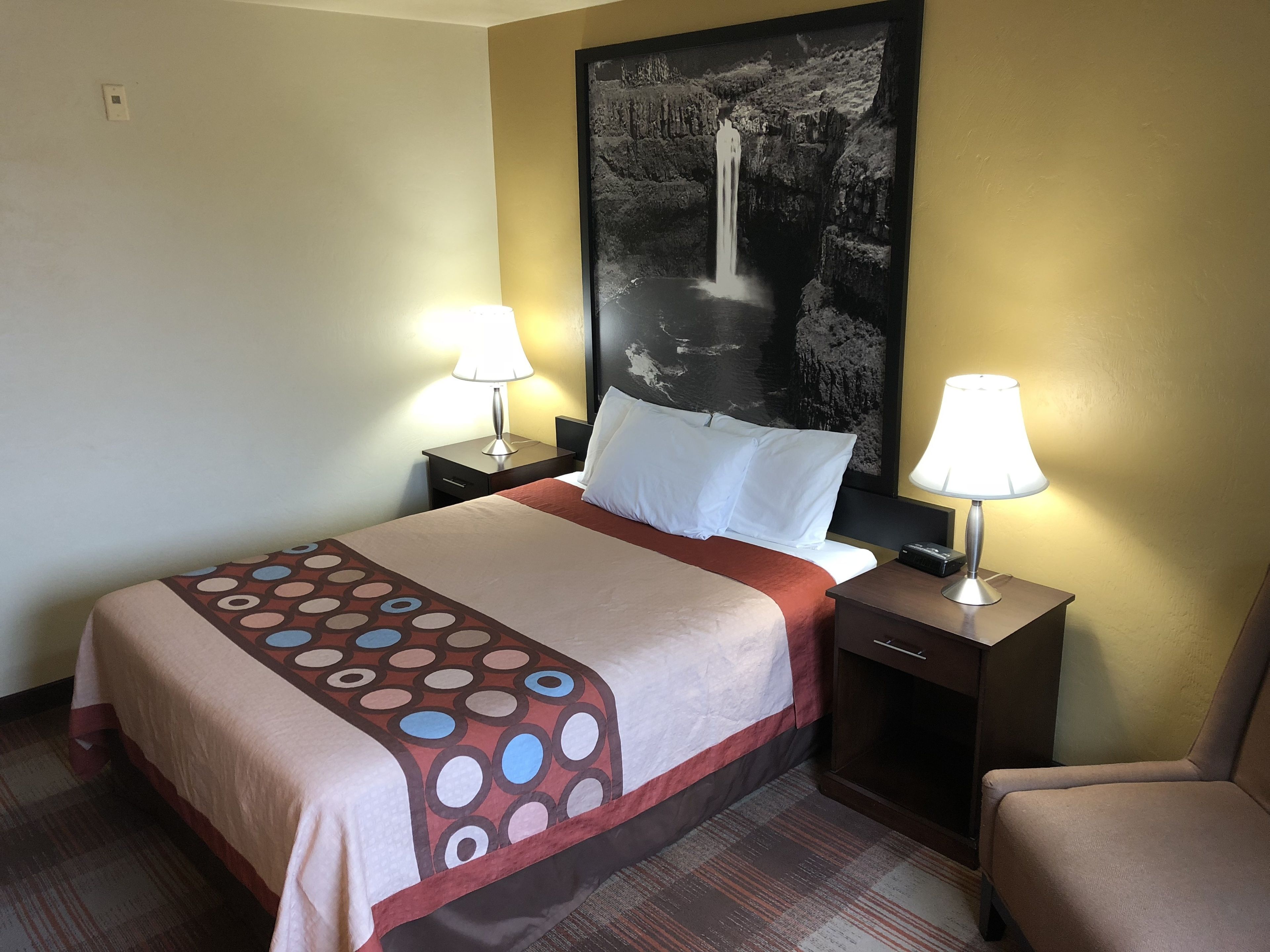 A hotel room with a bed, two bedside tables with lamps, a chair, a large abstract painting on the wall, and a patterned bedspread.