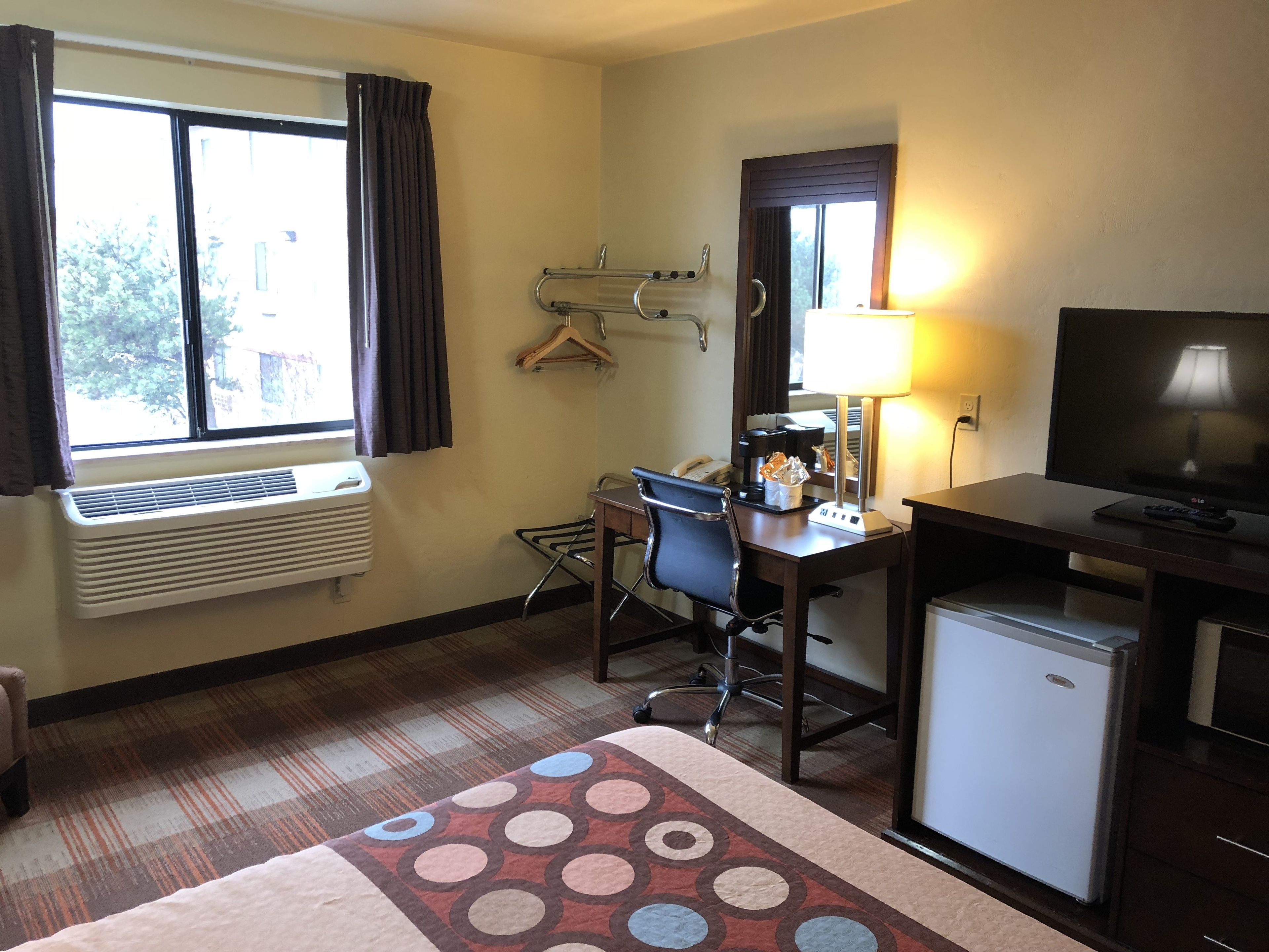 A hotel room with a bed, a desk with a chair, a television, a small refrigerator, a heater, a window with curtains, and a wall-mounted heater.