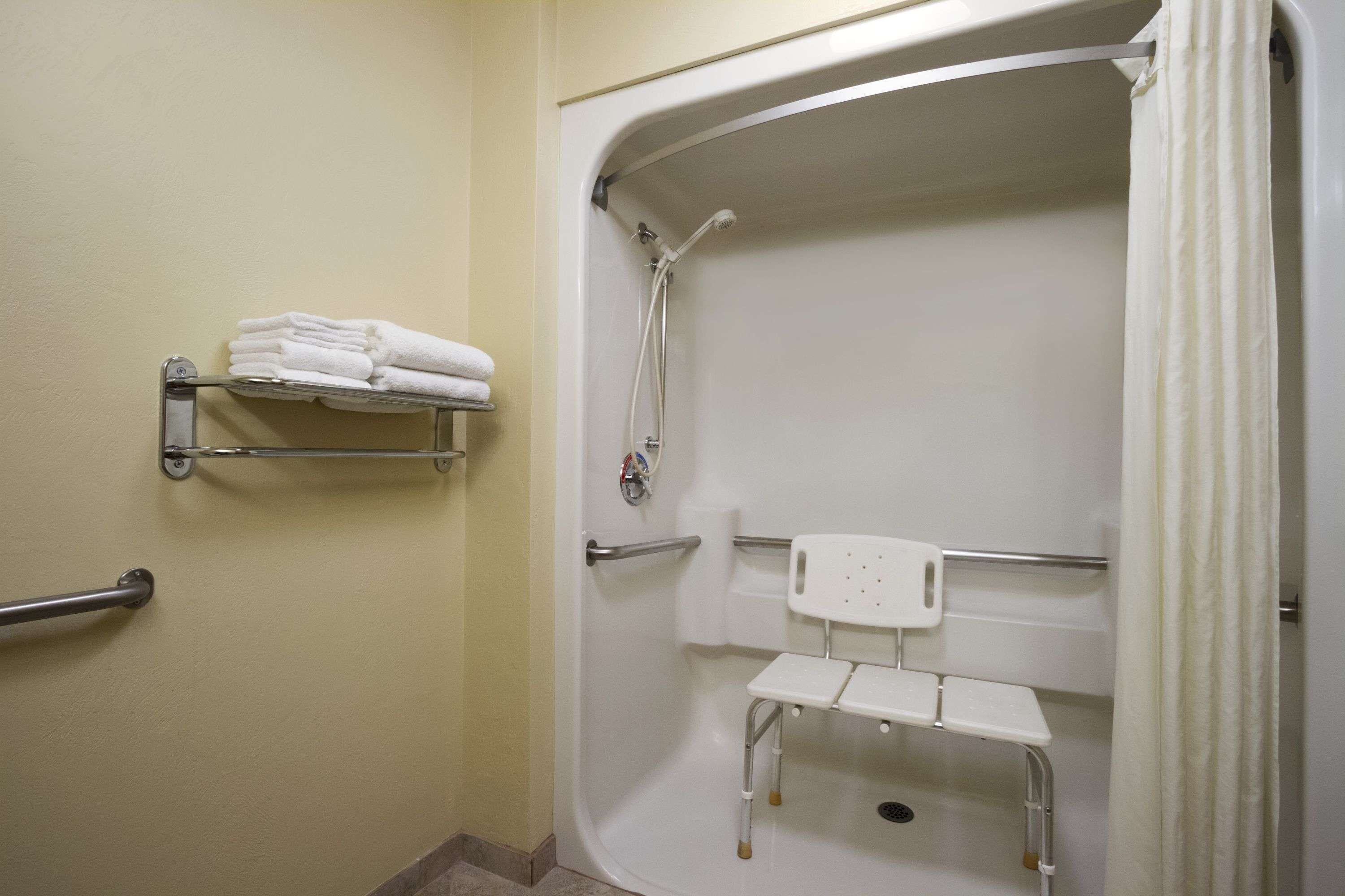 A bathroom with a shower, grab bars, a white chair, and a towel rack.