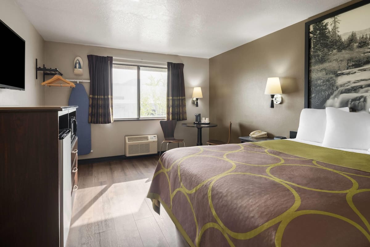A hotel room with a bed, a desk, a chair, a window with curtains, a wall-mounted TV, and a painting.