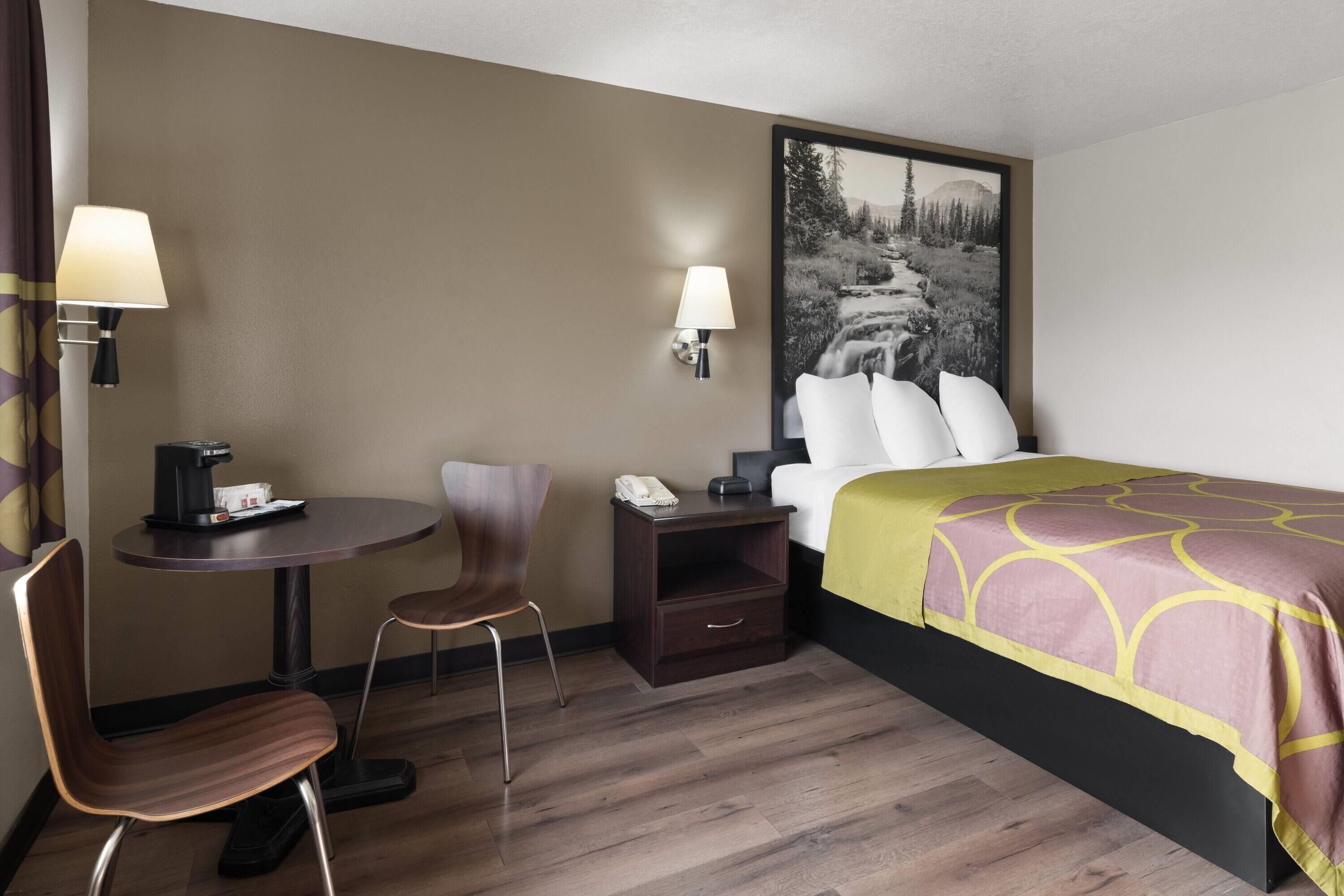 Una habitación de hotel con cama, un escritorio con lámpara, una silla, una mesita de noche con teléfono y una lámpara de pared.