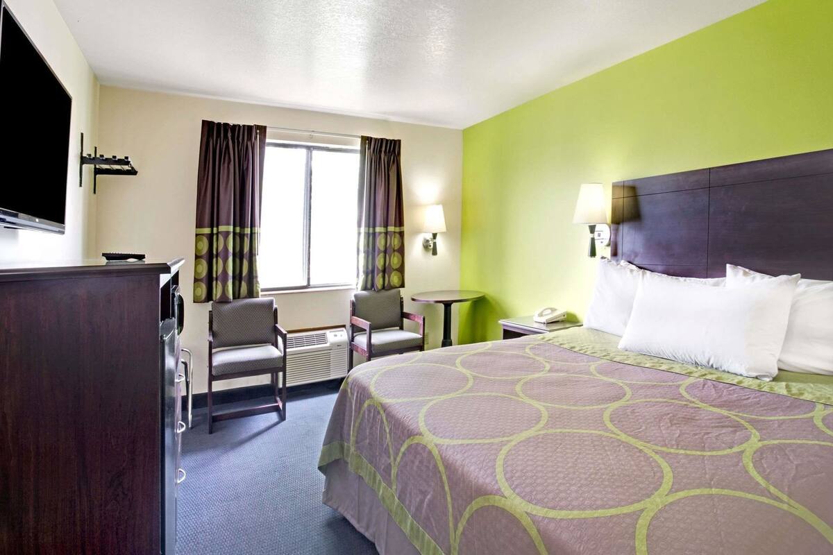 A hotel room with a bed, two chairs, a desk, a TV, and a window with curtains.