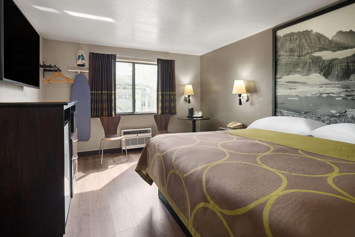A hotel room with a bed, a desk, a chair, a TV, a window with curtains, a lamp, and a painting.