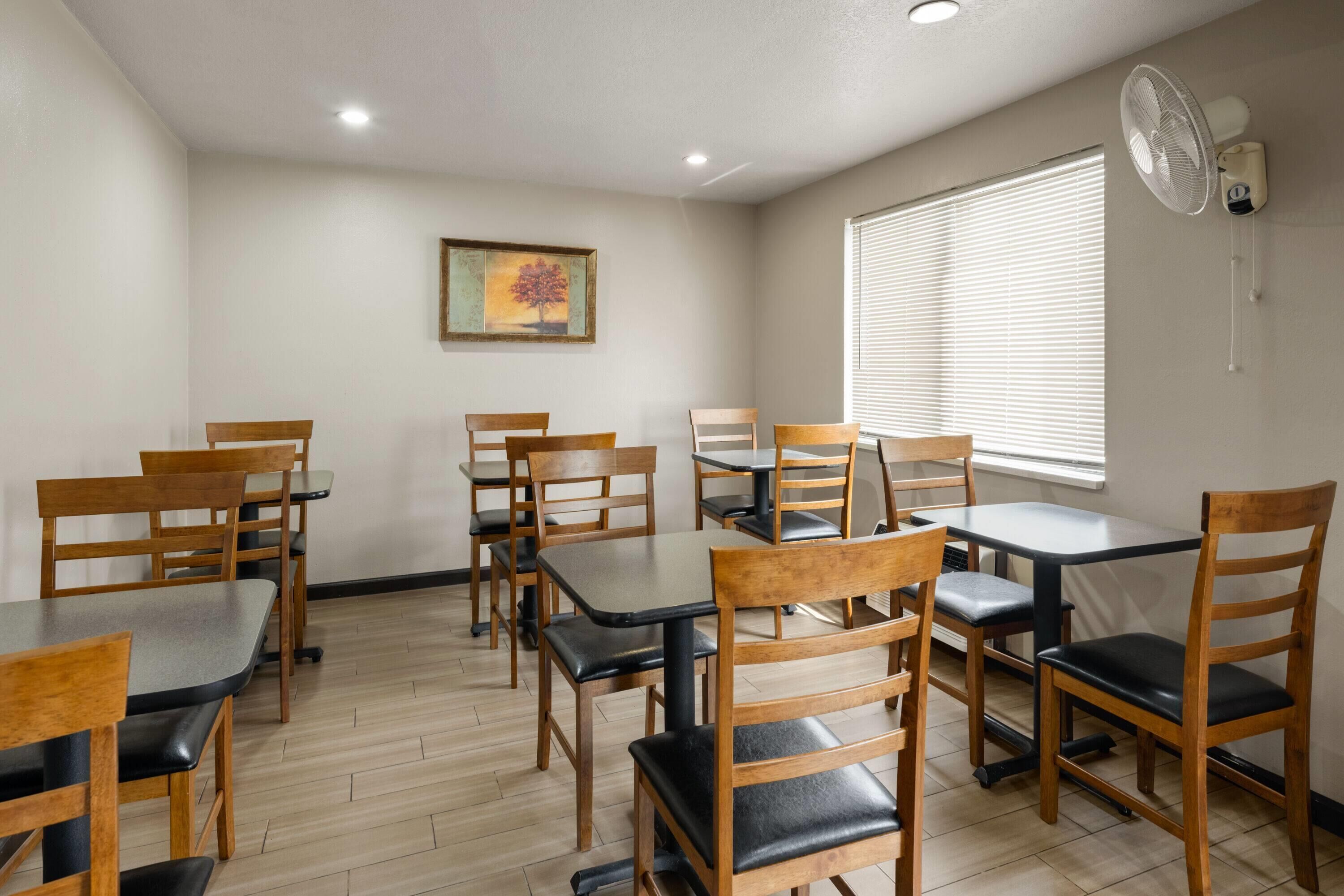 A room with wooden chairs and tables, a painting on the wall, and a window with blinds.