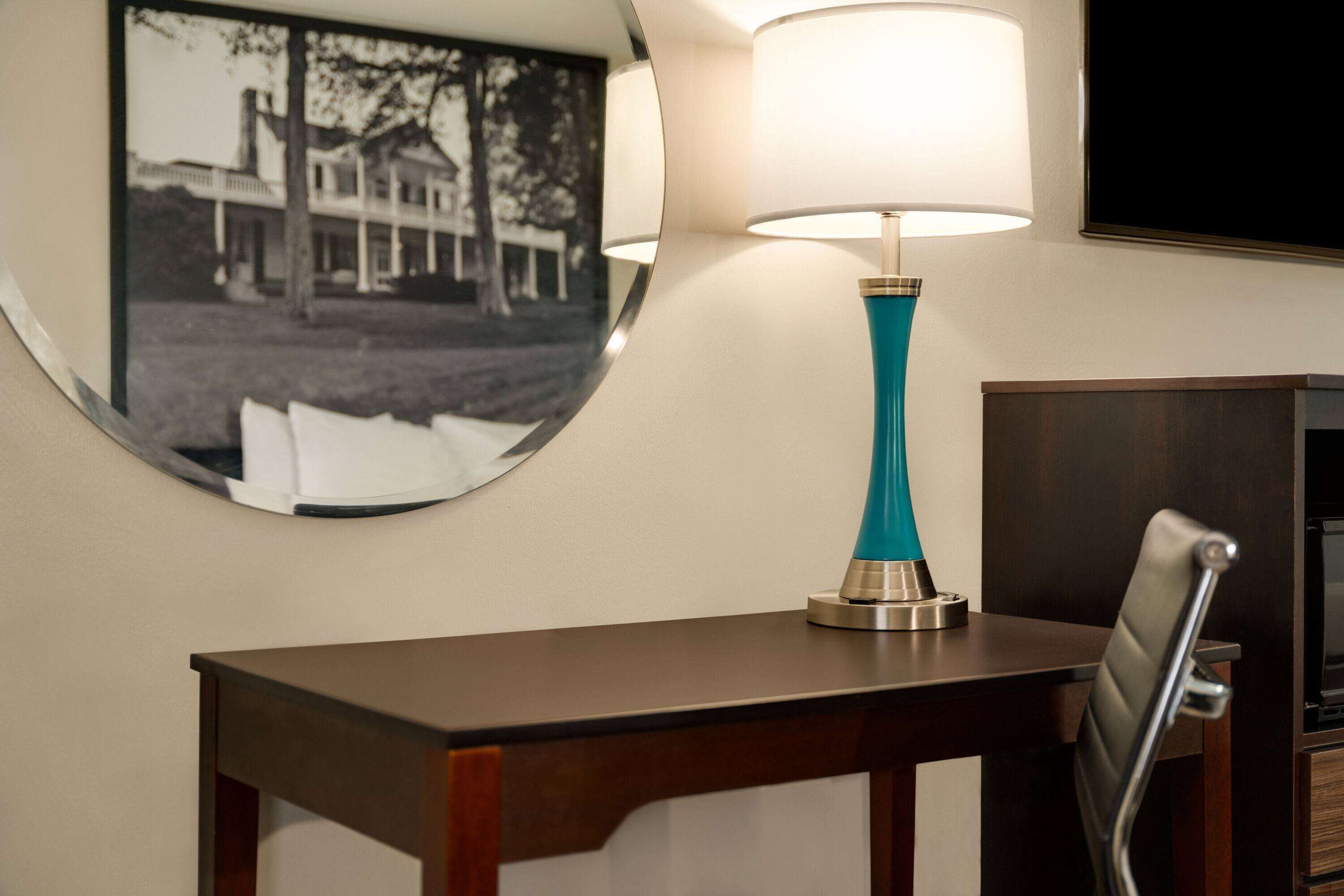A room with a wooden desk, a teal lamp, a chair, and a framed picture on the wall.