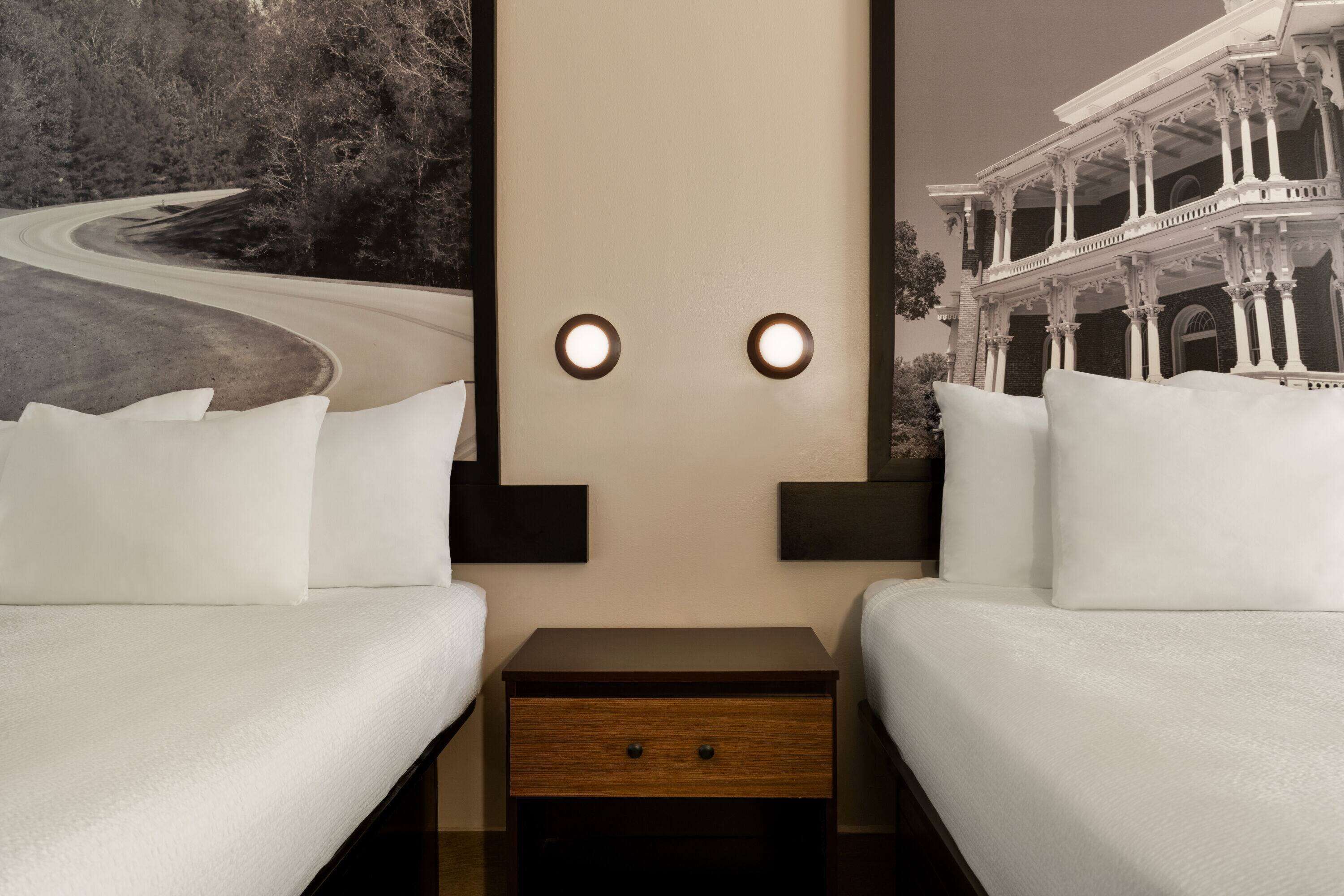 A hotel room with two beds, a nightstand, and a wall with a painting of a road and a building.