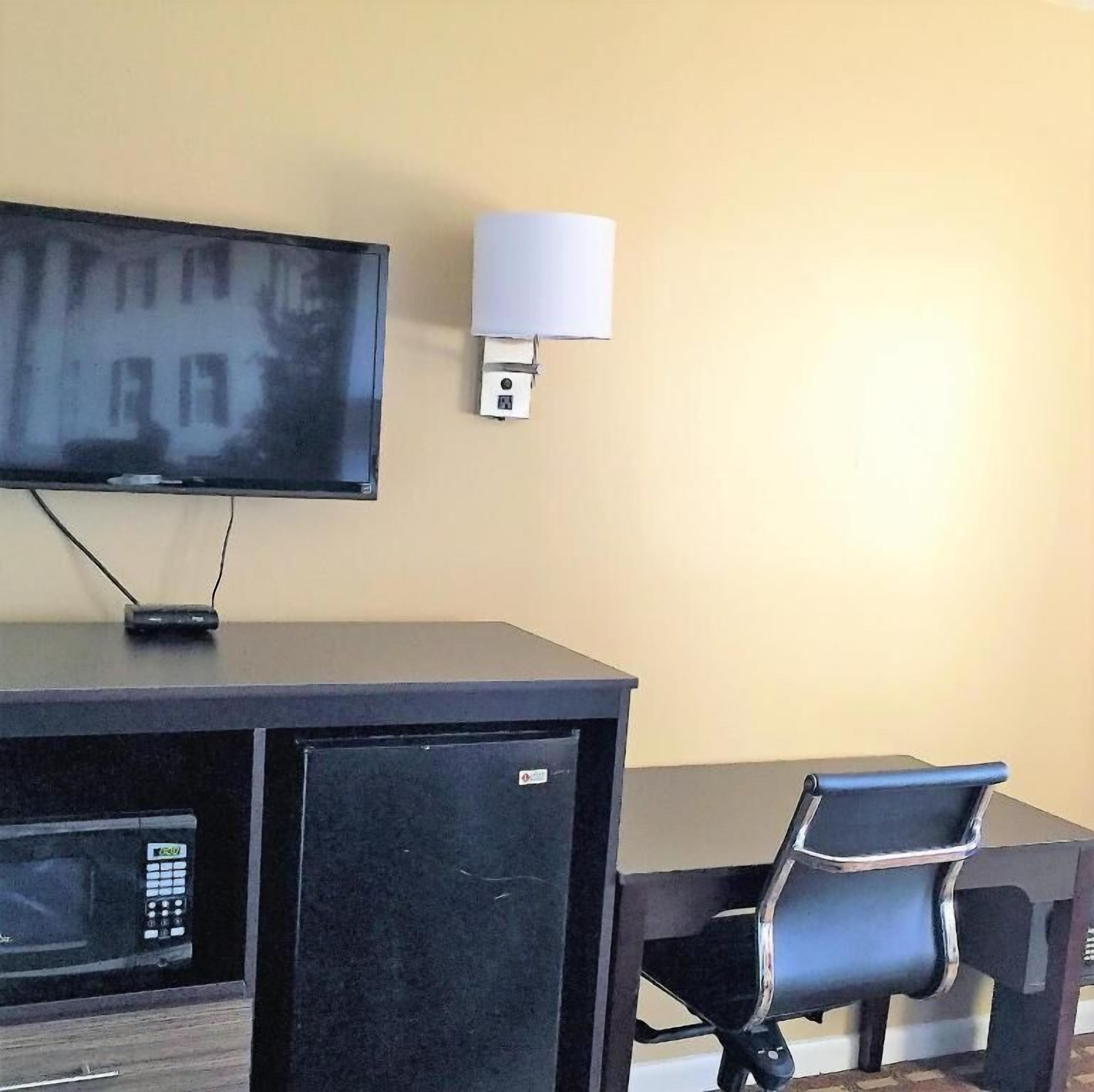 A room with a flat-screen TV mounted on a dark desk, a small refrigerator, a chair, and a wall-mounted lamp.