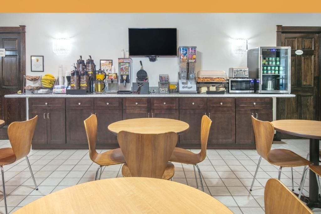 A modern kitchen with a bar area, equipped with coffee machines, a microwave, and a refrigerator. There are wooden chairs and a round table in the center.