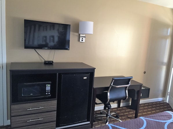 A compact hotel room with a flat-screen TV mounted on the wall, a small refrigerator, a desk with a chair, and a patterned rug on the floor.