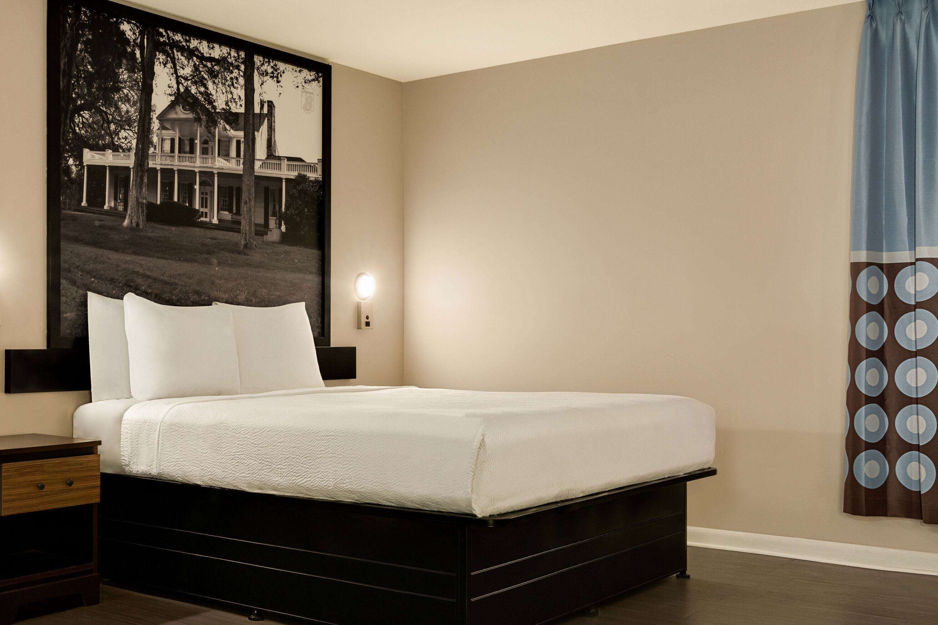 A bedroom with a large bed, a nightstand, a wall-mounted lamp, and a large framed picture of a house.