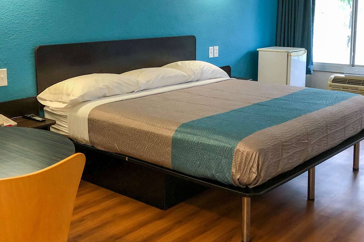 A single bed with a dark headboard, a bedside table, and a chair. There is a window with curtains, an air conditioning unit, and a small white appliance on the wall.