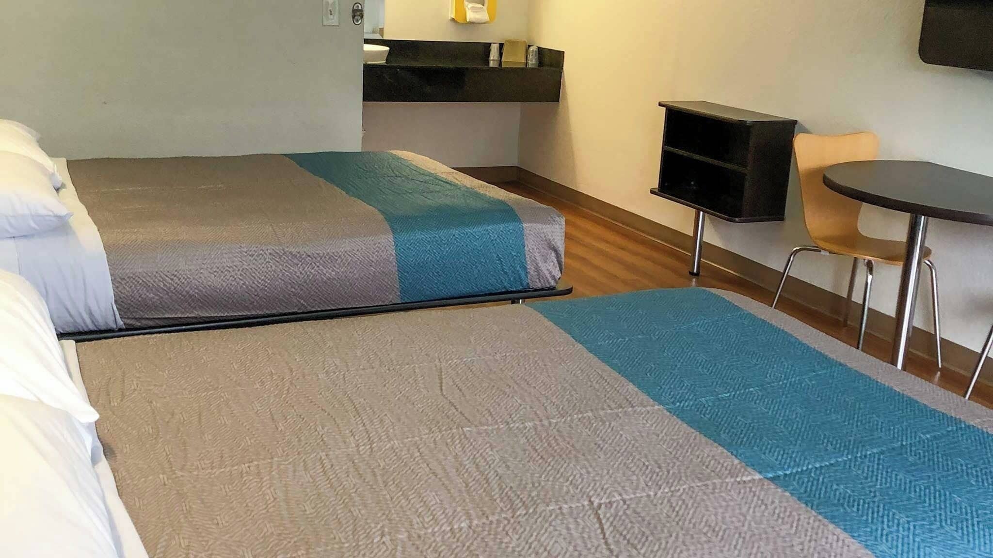 A hotel room with two beds, a small table, a chair, and a shelf with toiletries.
