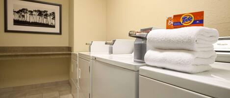 A hotel bathroom with a washing machine, a stack of white towels, and a Tide detergent box.