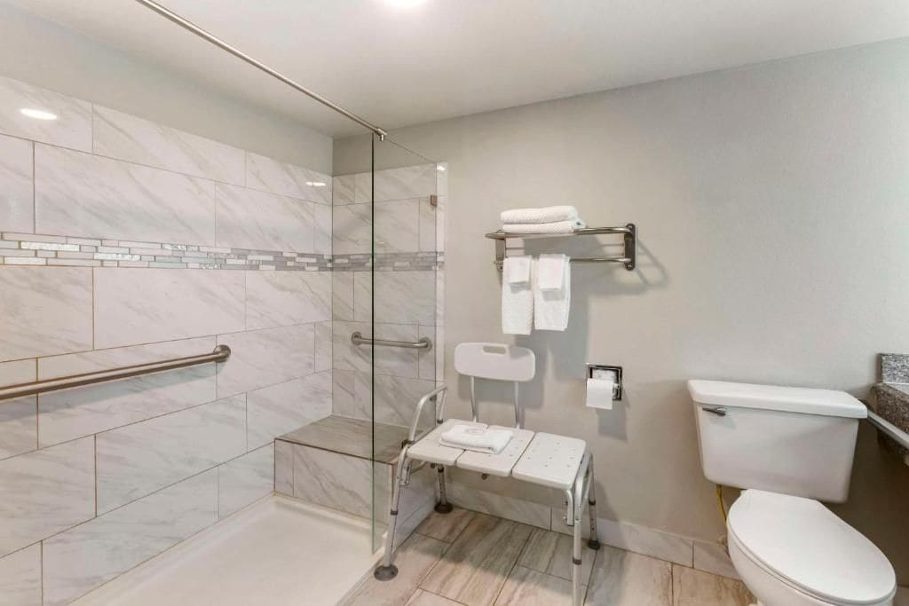 Un baño moderno con cabina de ducha de vidrio, un asiento accesible para silla de ruedas, un inodoro y un perchero para toallas.