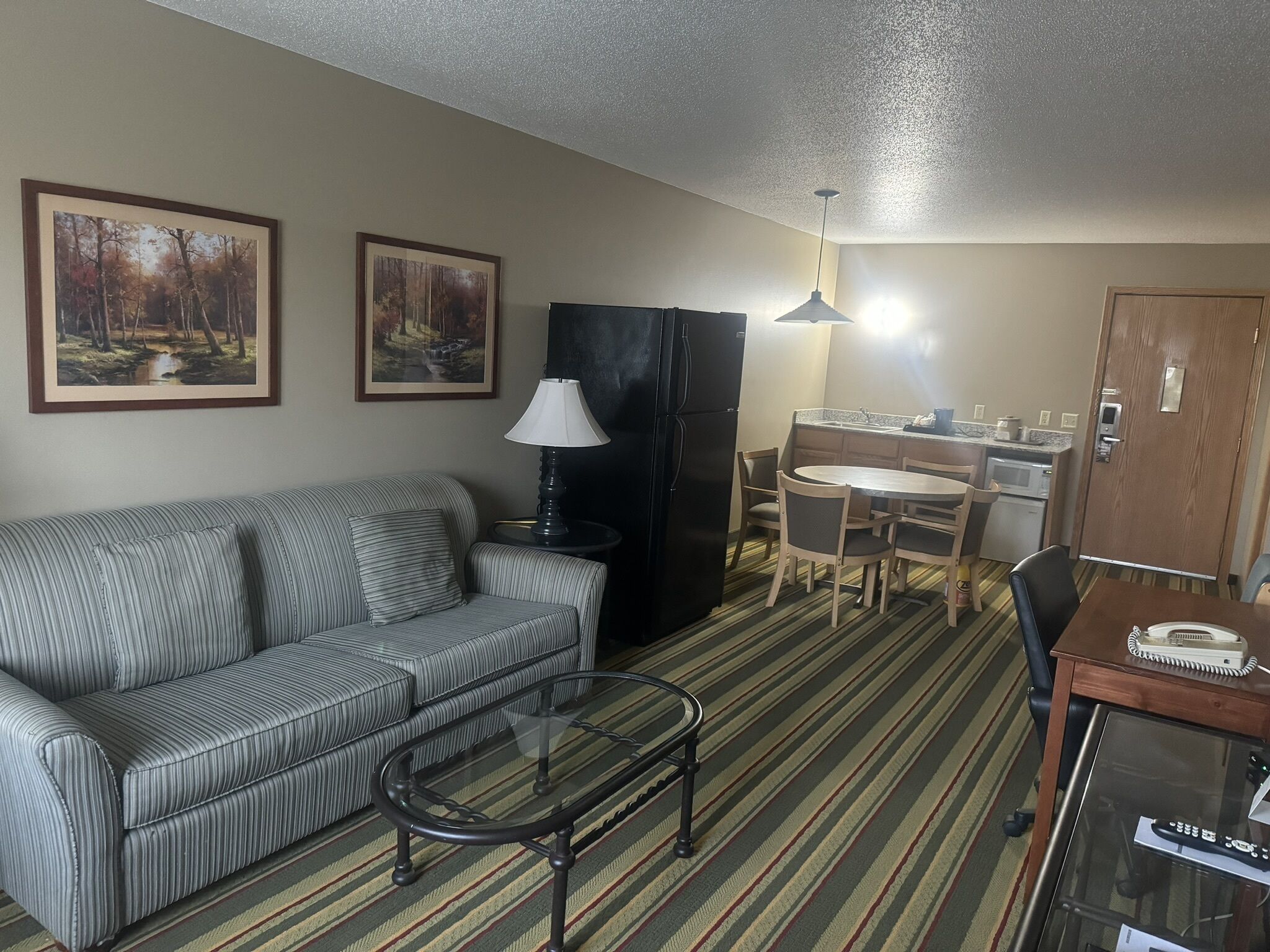 A compact living space with a sofa, a glass coffee table, a dining table with chairs, a kitchenette, and two framed pictures on the wall.