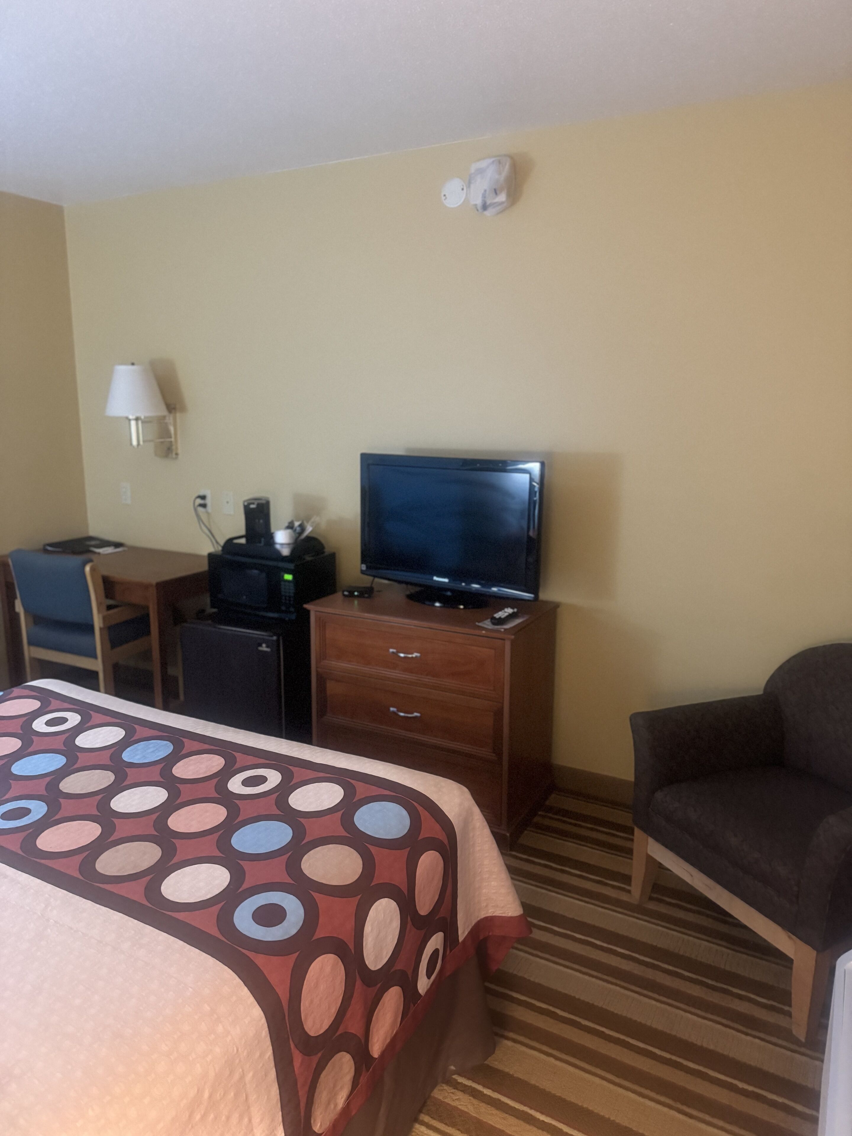 A hotel room with a bed, a desk with a computer, a TV on a dresser, a lamp, and a chair.