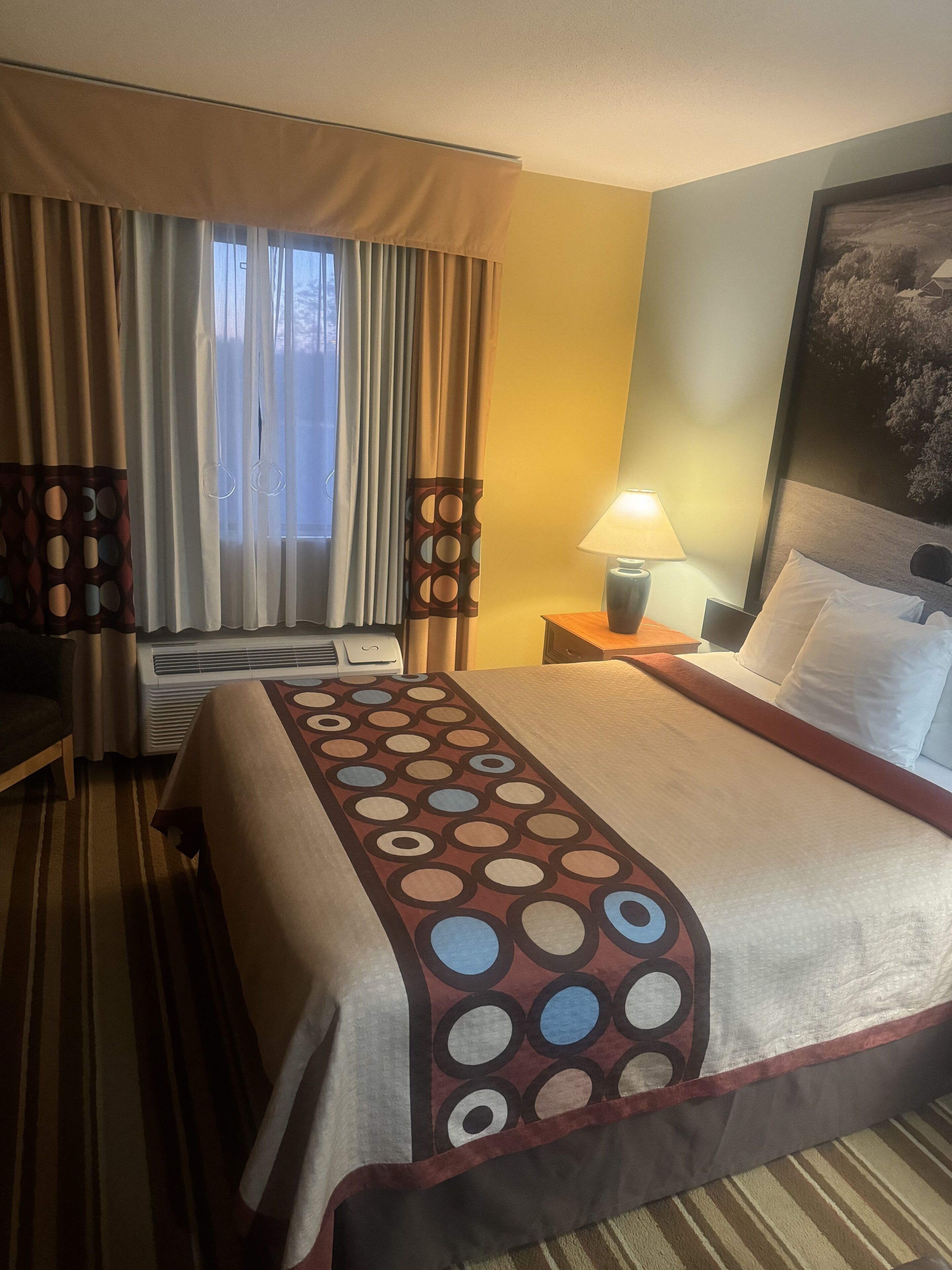 Una habitación de hotel con cama, escritorio, lámpara y ventana con cortinas.