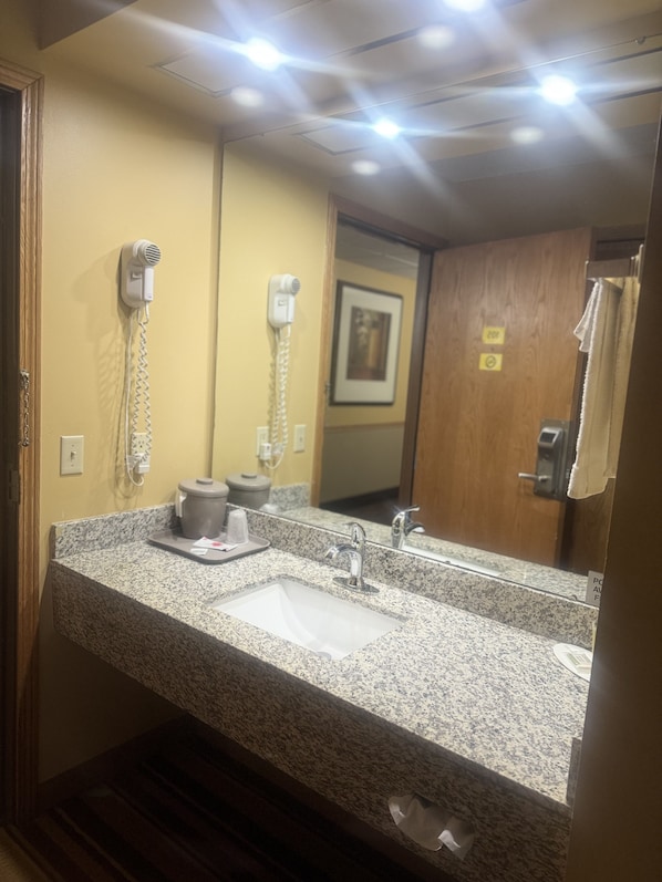 Un baño con lavamanos de encimera de granito, dos secadores de pelo montados en la pared, un cuadro en la pared y una puerta de madera.