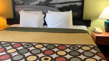 A hotel room with a bed, two pillows, a patterned throw, a nightstand with a lamp, and a framed landscape picture on the wall.