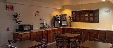 A compact kitchen with wooden cabinets, a microwave, a refrigerator, and a coffee machine. There are two wooden tables with chairs. An EXIT sign is visible on the wall.