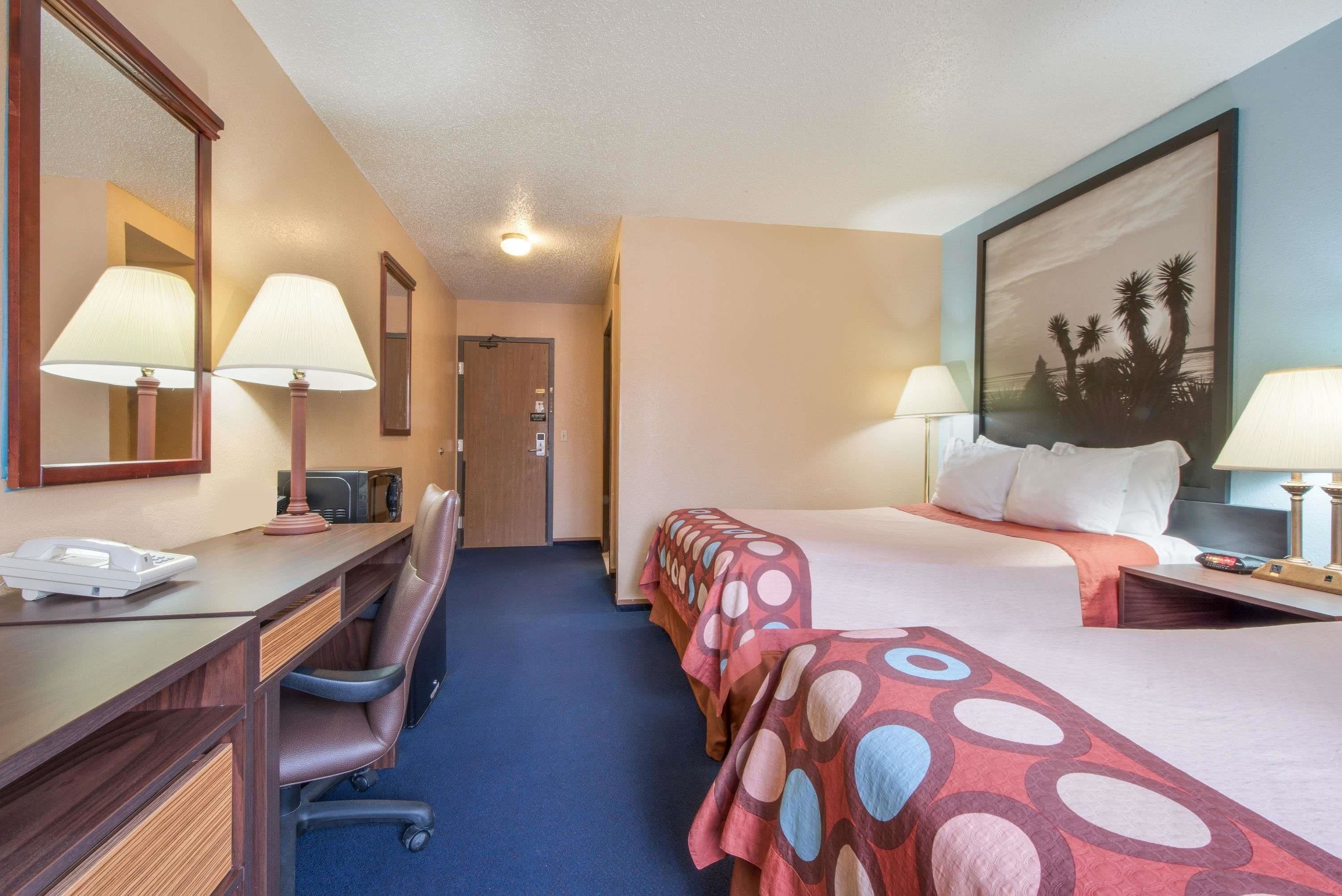 A hotel room with a desk, two beds, a chair, and a large painting on the wall.