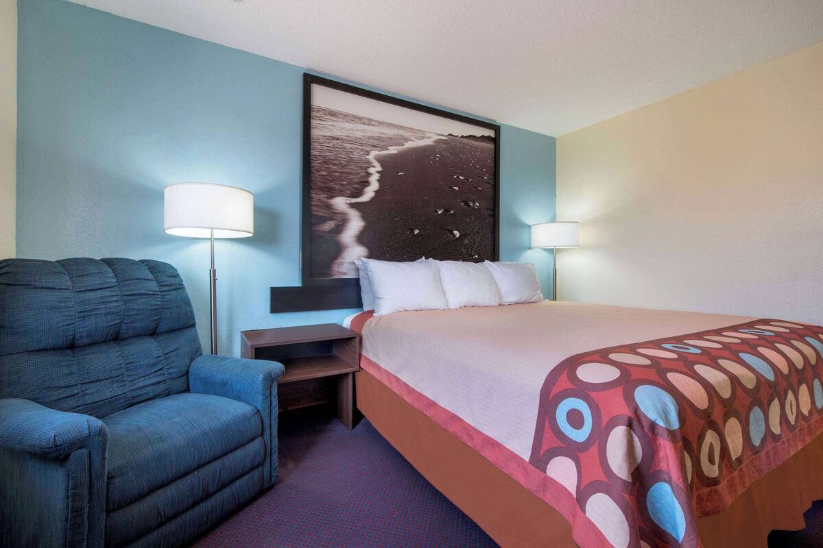 Ein Hotelzimmer mit einem Bett, einem Sessel, zwei Nachttischlampen und einem großen Bild einer Strandansicht in einem Rahmen.