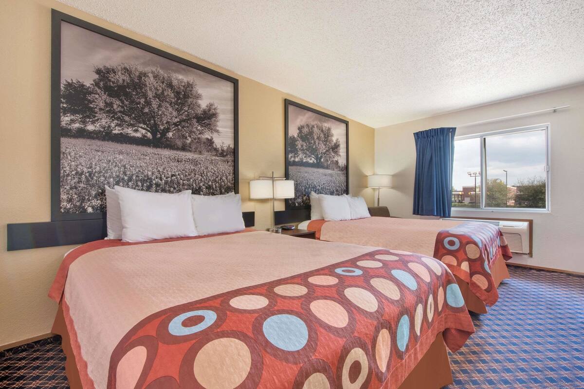 Ein Hotelzimmer mit zwei Betten, einem großen Fenster mit Vorhängen und einem an der Wand hängenden Bild.