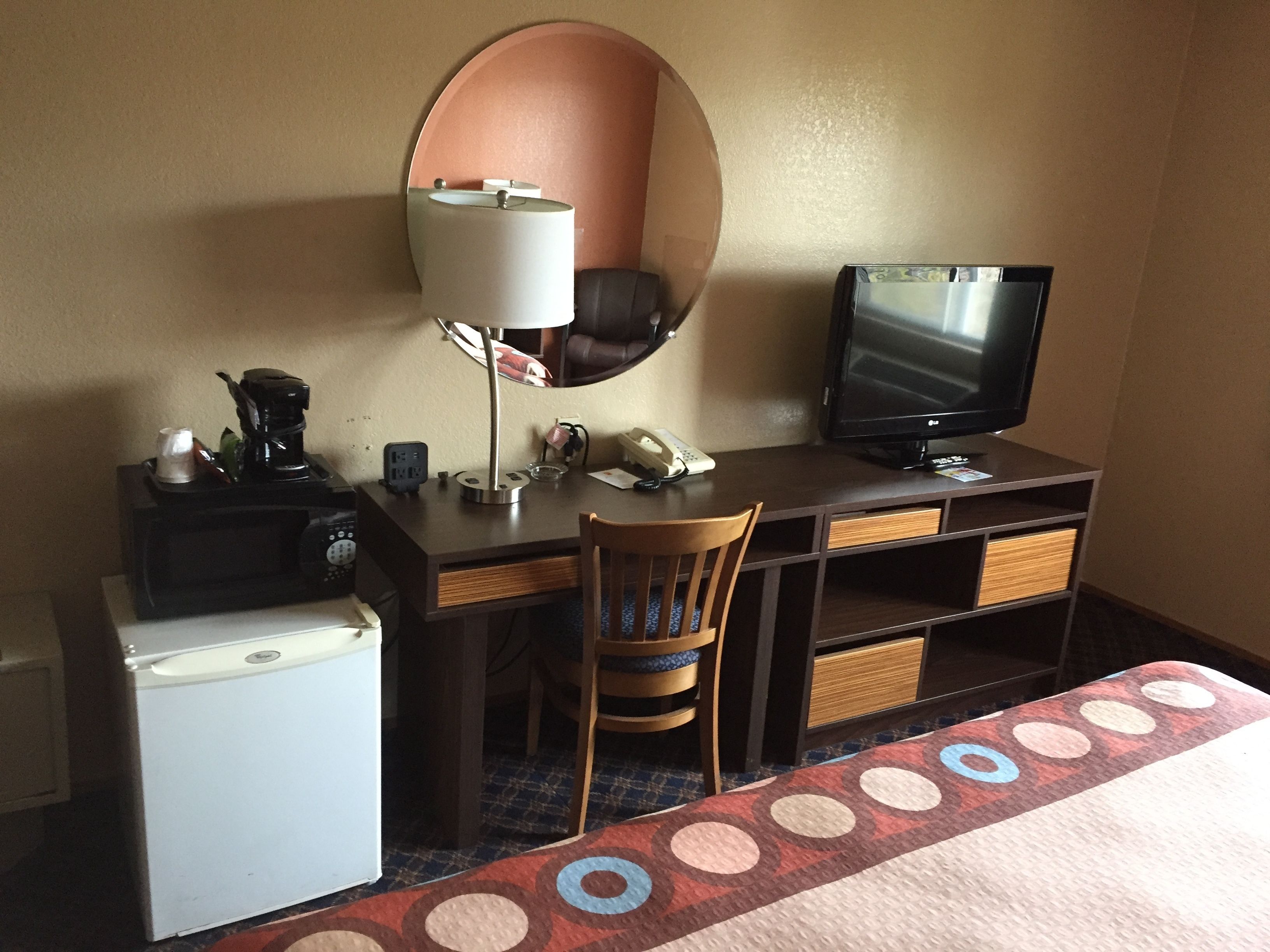 A hotel room with a desk, a small refrigerator, a chair, a bed, a television, and a mirror.