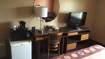 A hotel room with a desk, a small refrigerator, a chair, a bed, a television, and a mirror.