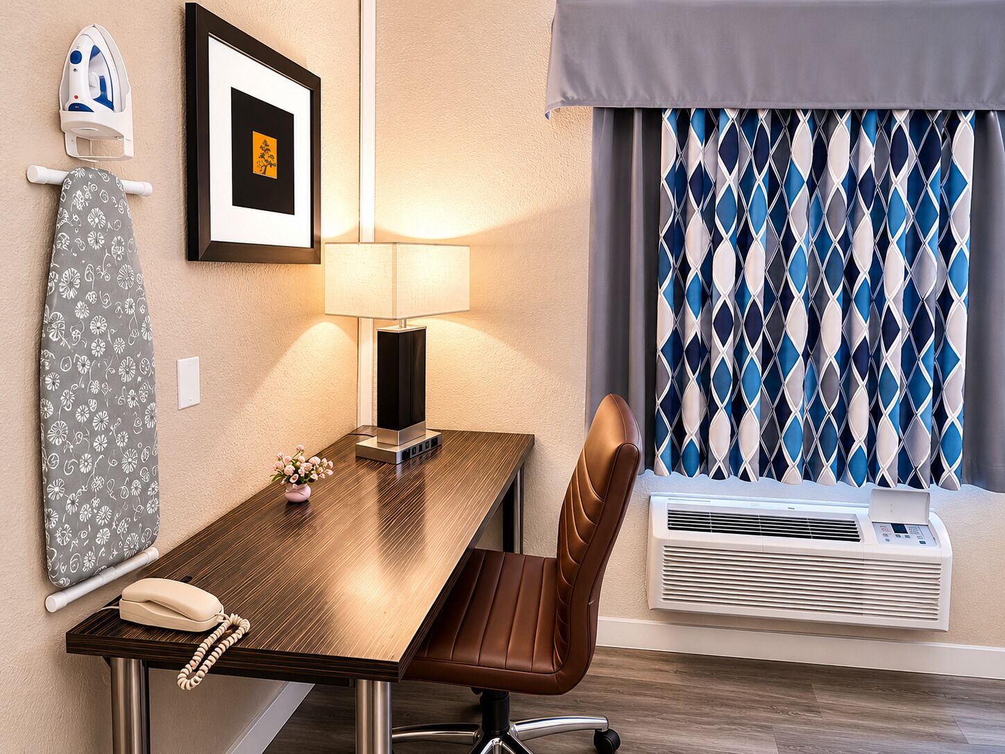 A hotel room with a wooden desk, a brown leather chair, a framed picture, a lamp, a telephone, an iron, a window with patterned curtains, and an air conditioning unit.