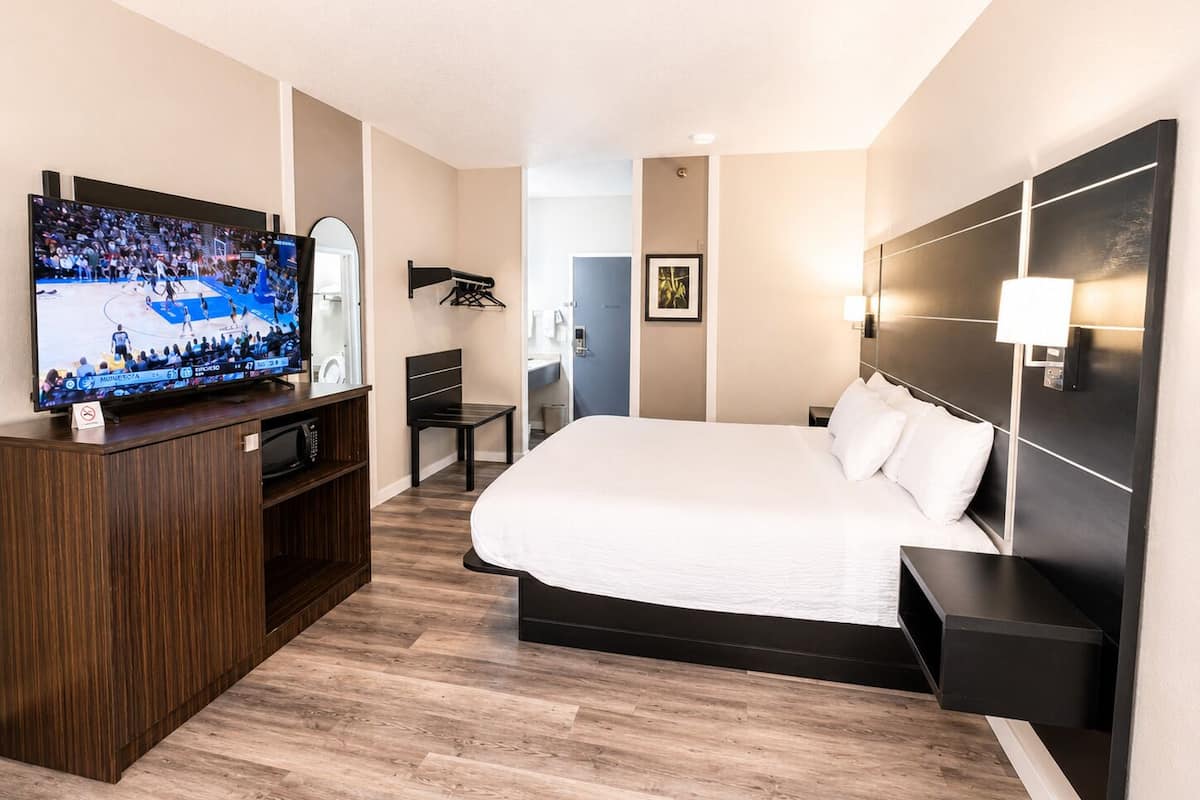 Habitación de hotel con una cama grande, un televisor sobre un escritorio de madera, una silla, una mesita pequeña y una lámpara de pared.