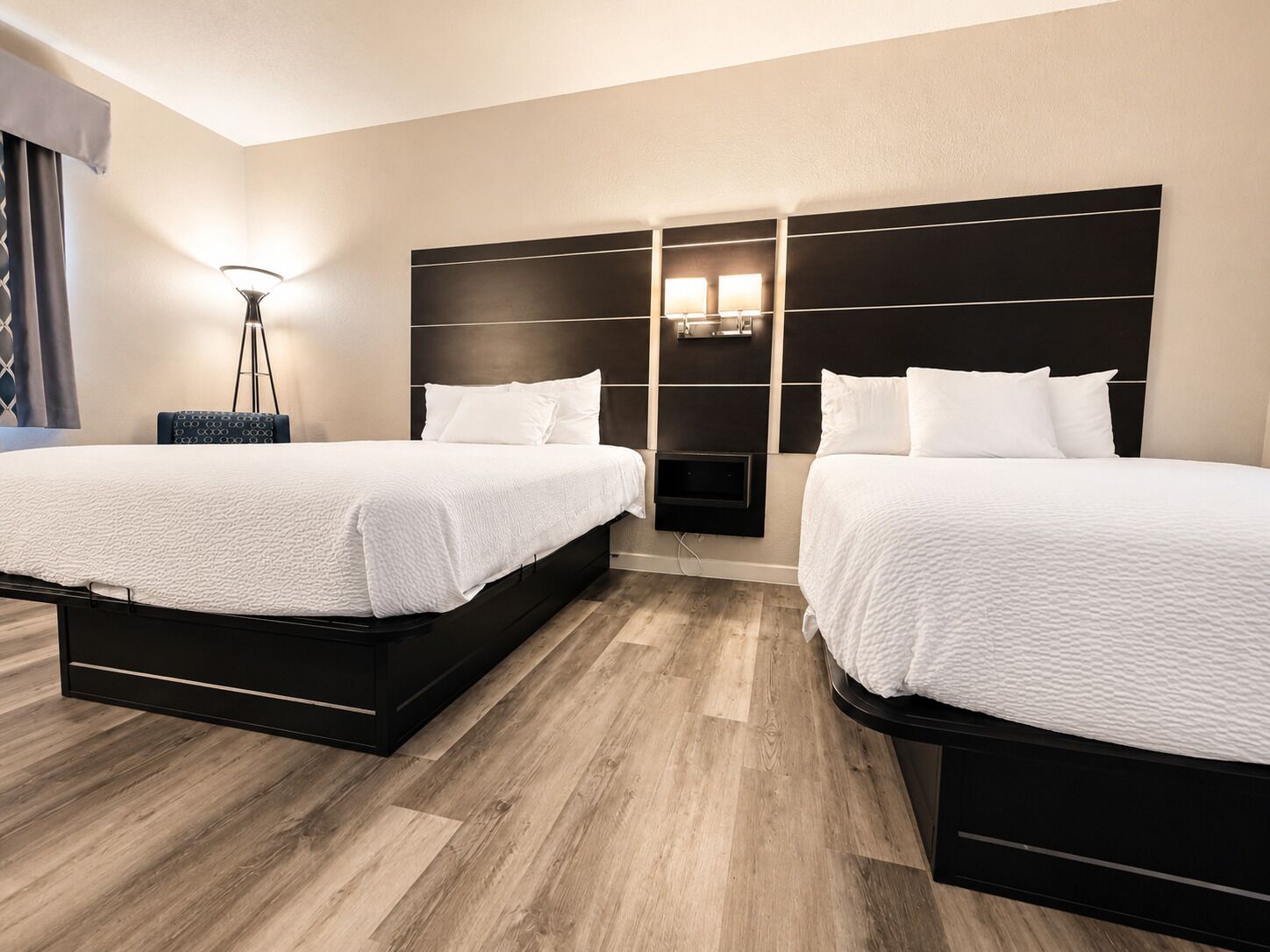 A hotel room with two beds, a bedside table, a lamp, and a wall-mounted light fixture.