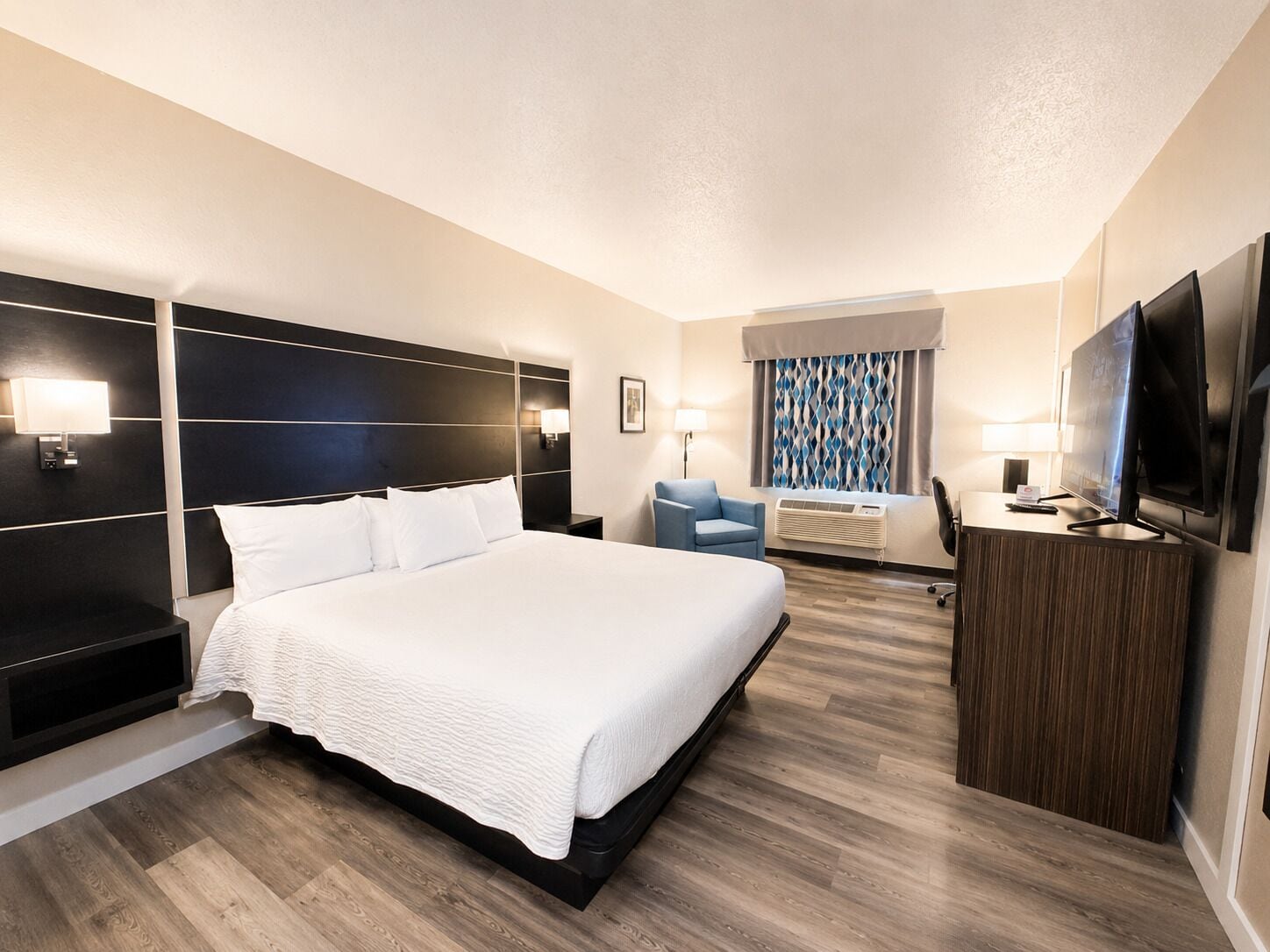A hotel room with a large bed, a desk with a TV, a chair, and a window with curtains.