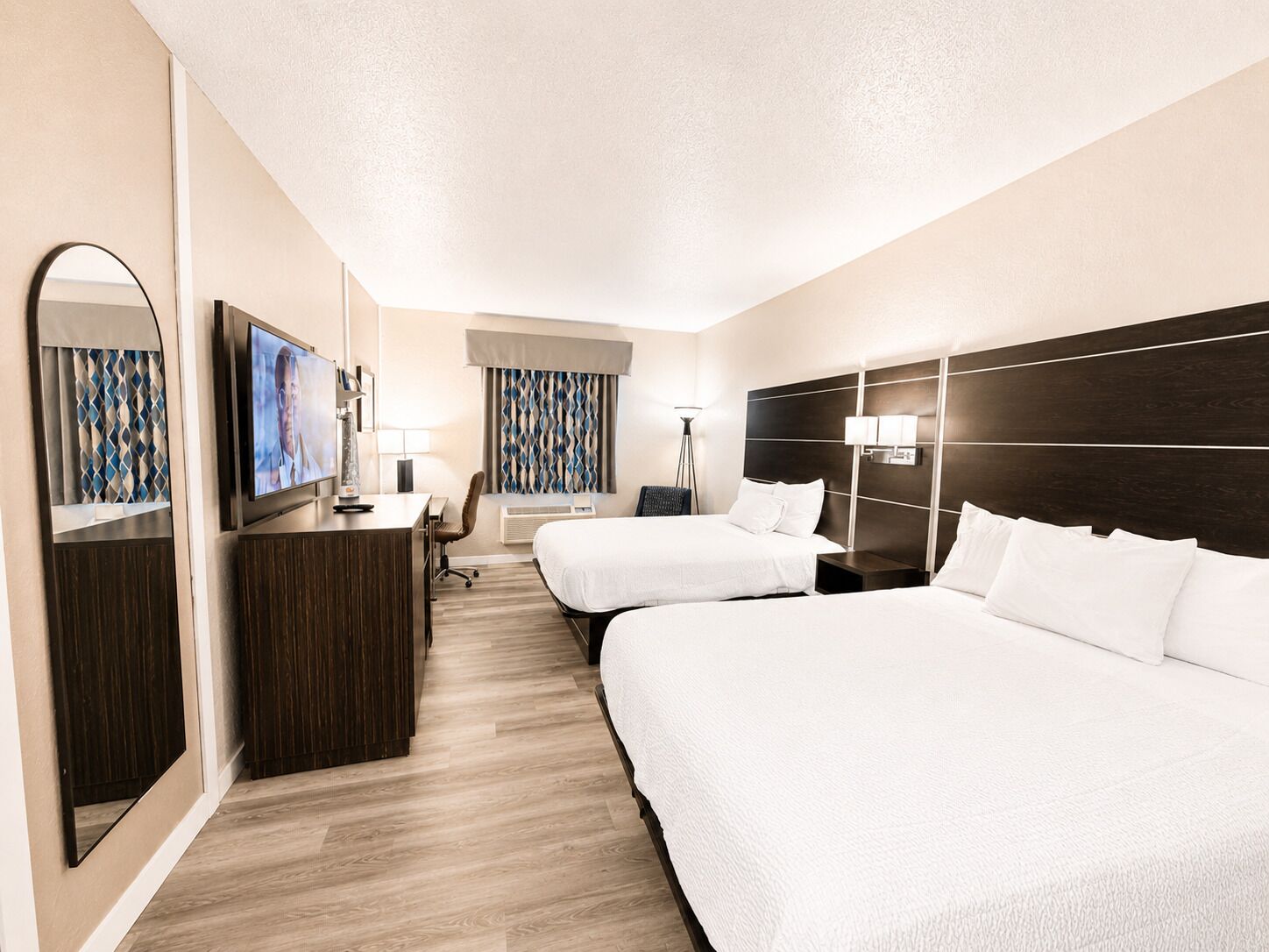 Habitación de hotel con dos camas, un escritorio, una silla, un televisor y un espejo grande.