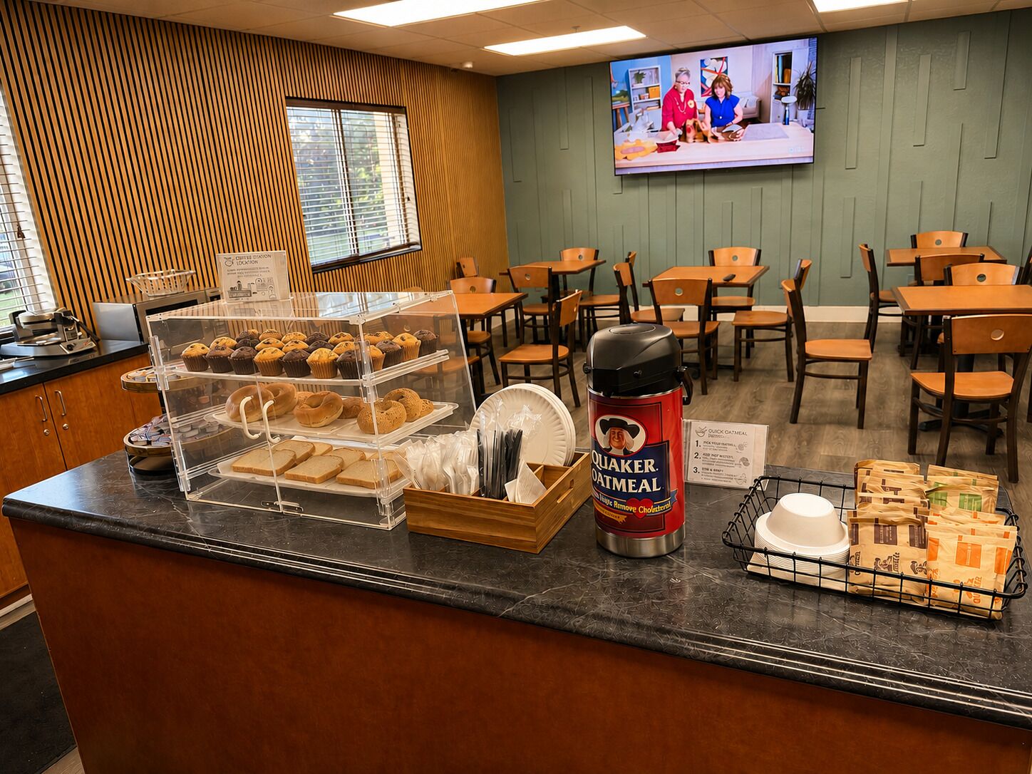 A breakfast area with a display case of pastries, a Quaker Oatmeal coffee maker, and a TV screen in the background.