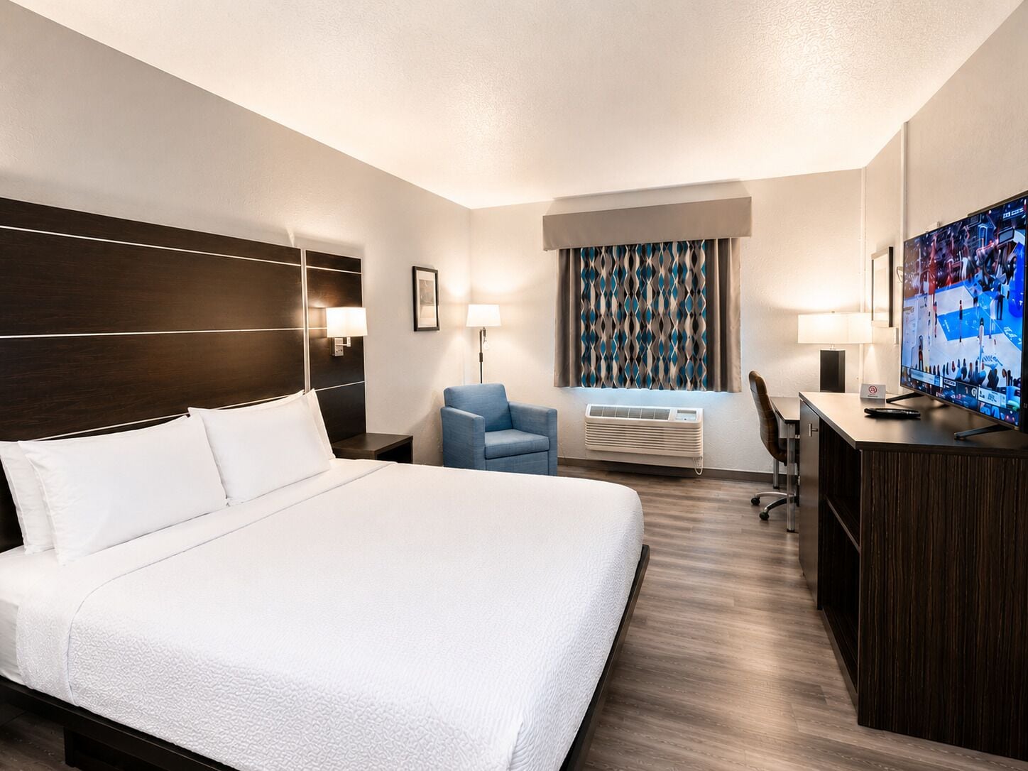 A hotel room with a large bed, a desk with a television, a chair, and a window with patterned curtains.