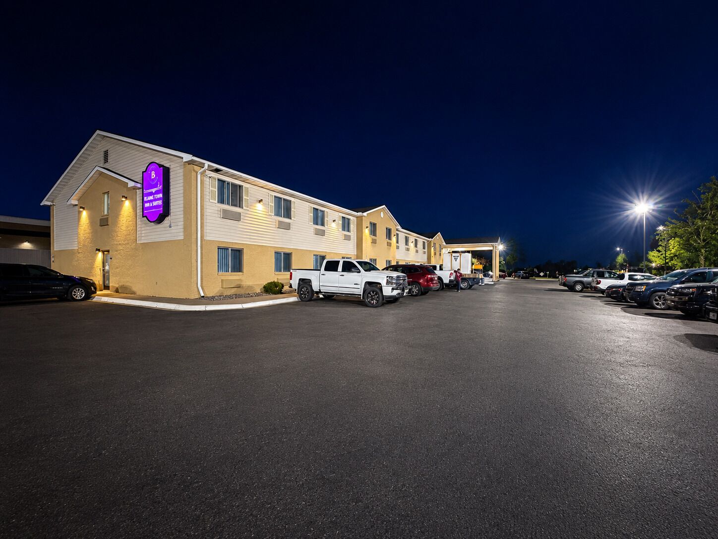 Un motel doté d’un parking, où plusieurs véhicules sont stationnés, et dont la façade est bien éclairée.