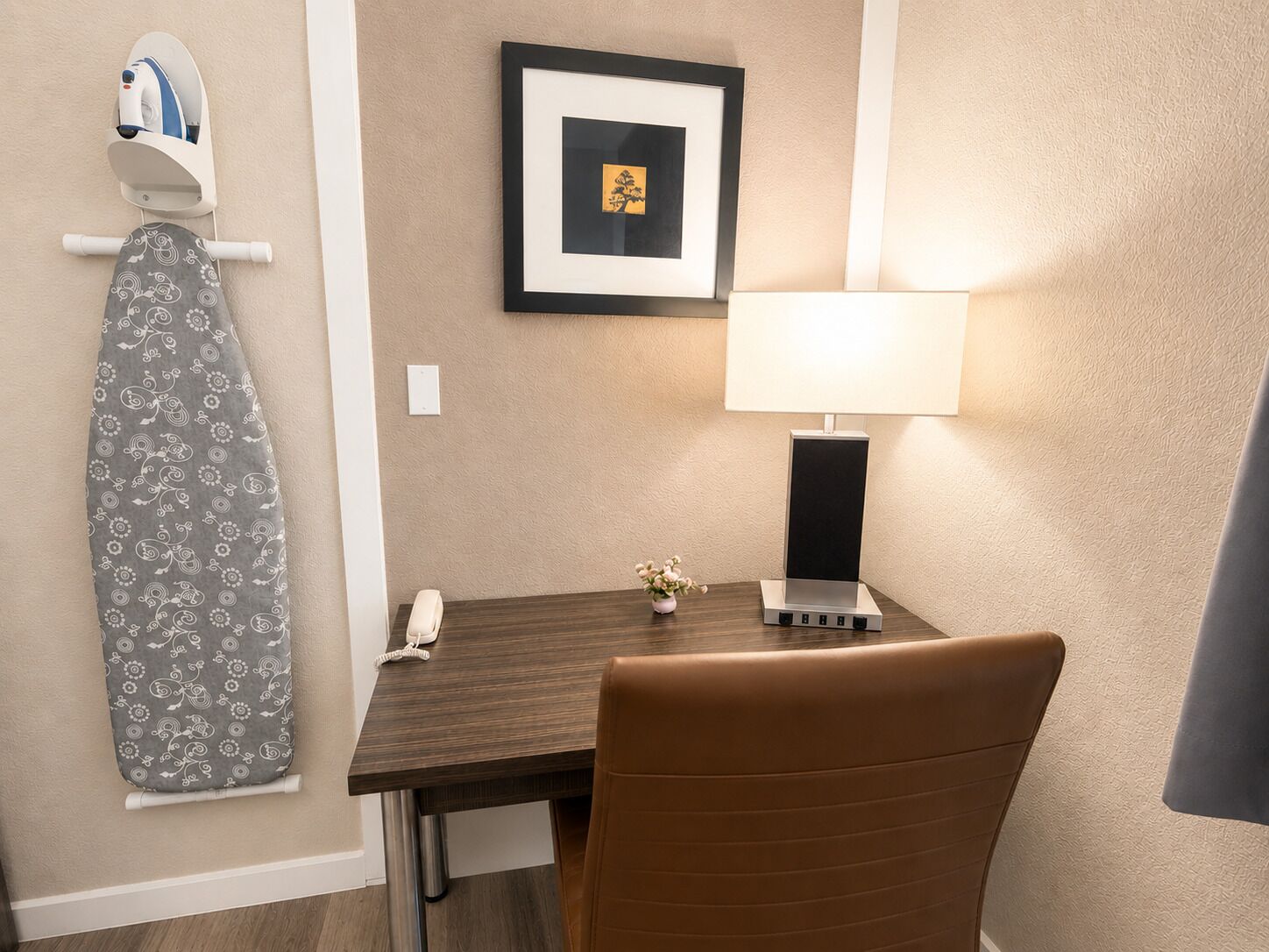 A hotel room with a wooden table, a brown leather chair, a framed picture on the wall, a wall-mounted ironing board, and a desk lamp.
