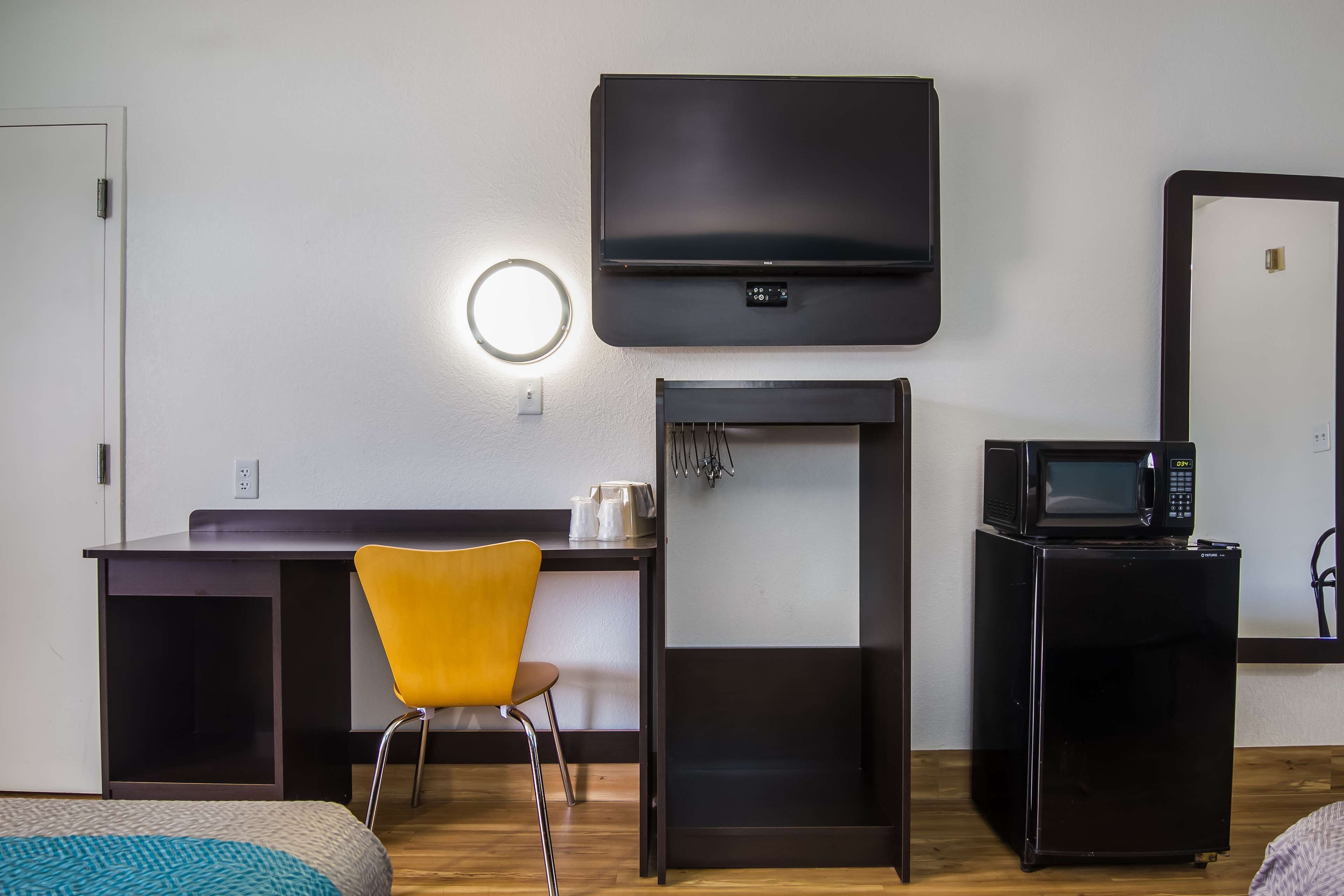 A compact hotel room with a bed, a desk with a chair, a TV mounted on the wall, a microwave, and a small refrigerator.