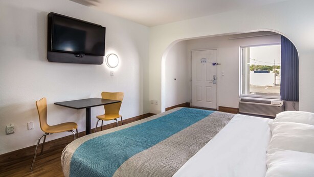 A hotel room with a bed, a desk with two chairs, a television mounted on the wall, and a door leading to a balcony.