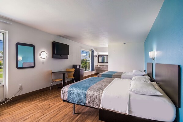 Una habitación de hotel moderna con suelo de madera, un escritorio con una silla, una cama con manta azul y blanca a rayas, un televisor de pantalla plana montado en la pared y una ventana con vistas a zonas verdes.