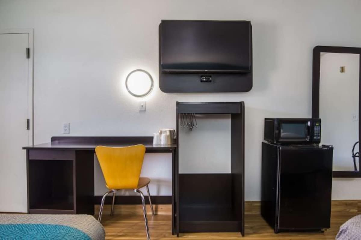 A hotel room with a bed, a desk with a chair, a TV mounted on the wall, a microwave, and a refrigerator.