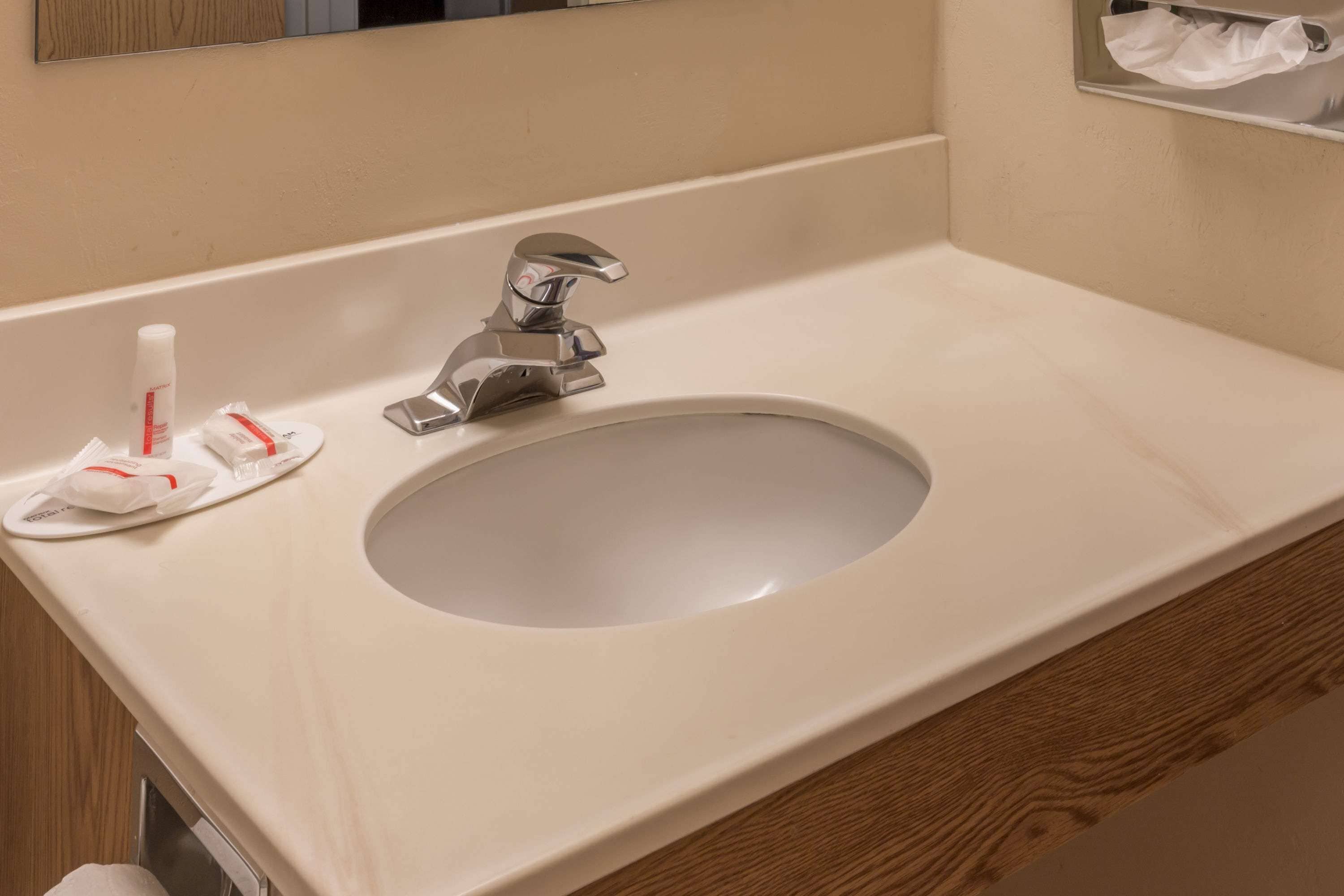 A bathroom sink with a mirror above it, a faucet, and a towel dispenser with a bottle and a small item on the counter.