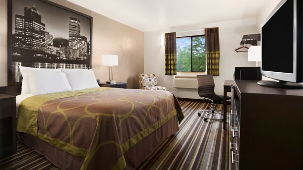 Habitación de hotel con una cama grande, un escritorio con computadora, una silla, una ventana con cortinas y un cuadro de un paisaje urbano en la pared.